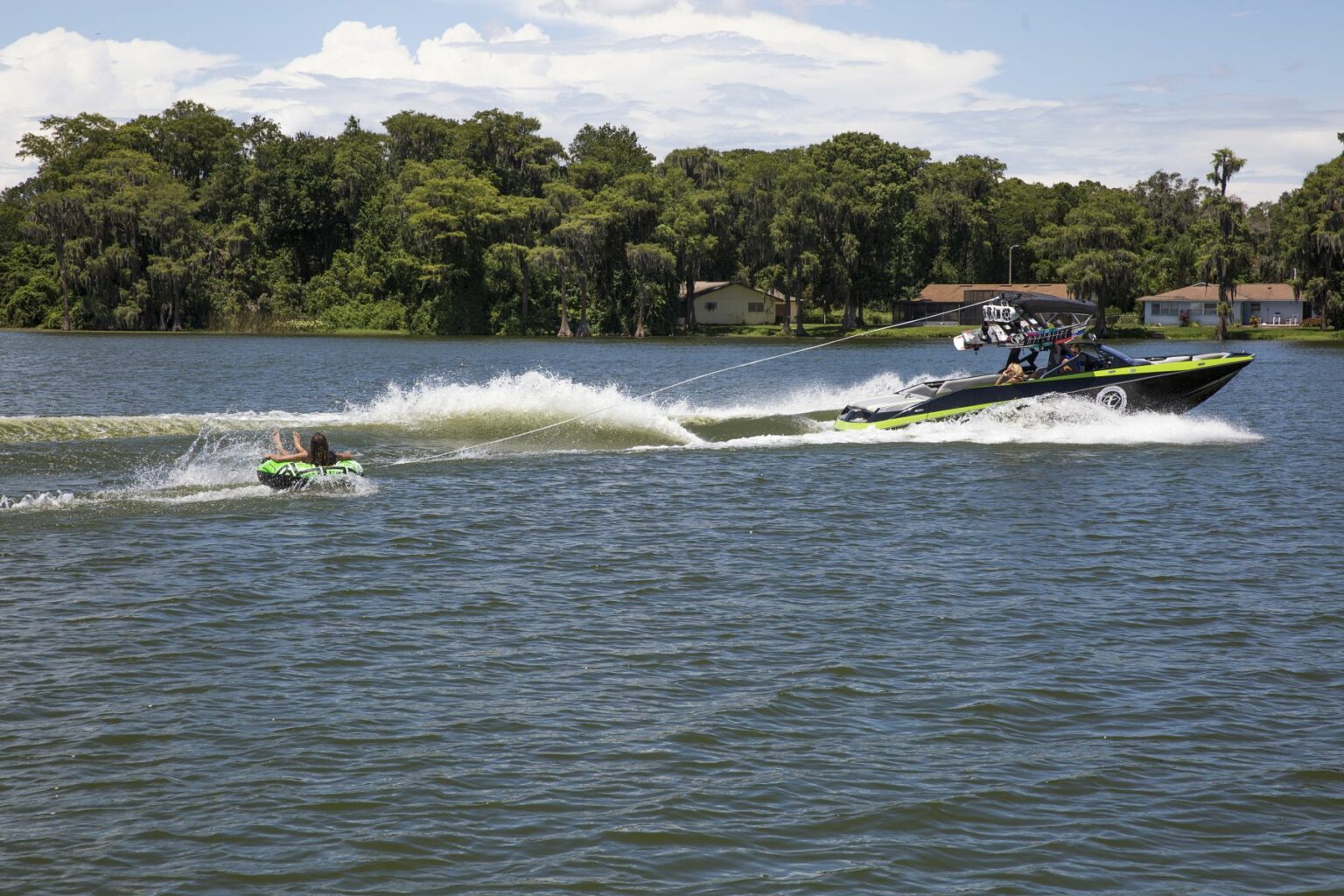 License-free image - A speedboat with several passengers tows a person on a green and yellow inflatable tube across a lake. The water is choppy from the boat&#039;s wake, and lush green trees line the distant shoreline under a partly cloudy sky. Tubing behind a wakeboard boat on Clear Lake in Orlando, Florida.