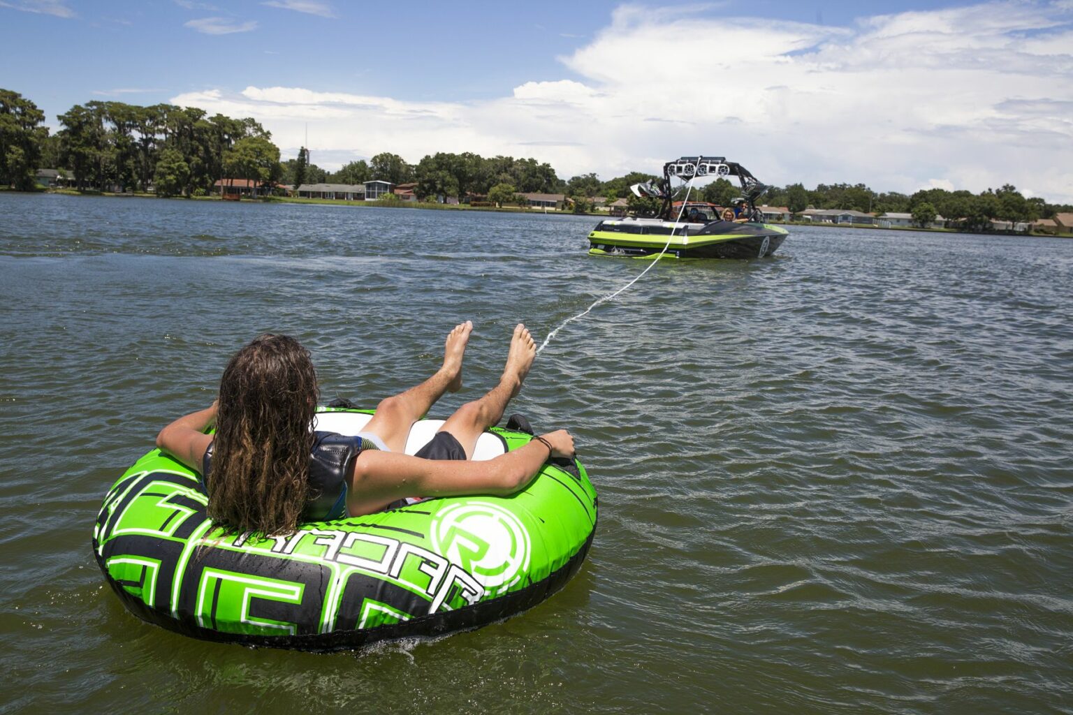 License-free image - A person with long hair sits in a green and black inflatable tube, being towed by a green and black boat on a lake. The sky is partly cloudy, and there are trees and buildings along the shoreline in the background. Tubing behind a wakeboard boat on Clear Lake in Orlando, Florida.