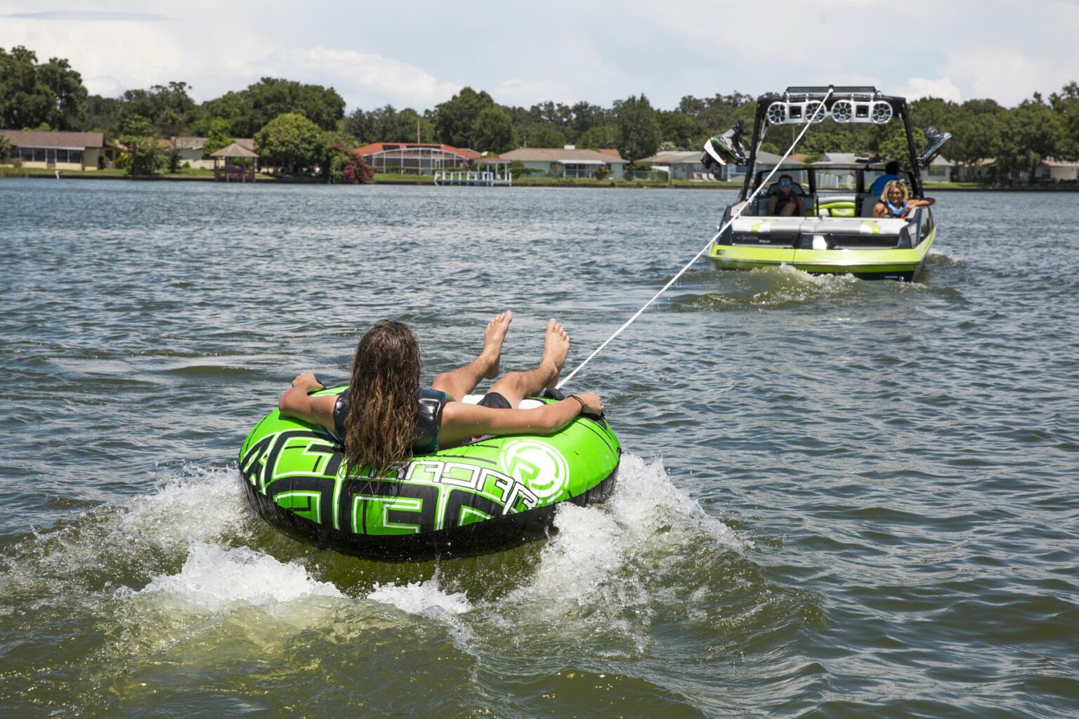 License-free image - A person on a green and black inflatable tube is being towed by a motorboat on a lake with tree-lined shores and houses in the background. The person is lying on their back with feet up, enjoying the water activity under a partly cloudy sky. Tubing behind a wakeboard boat on Clear Lake in Orlando, Florida.