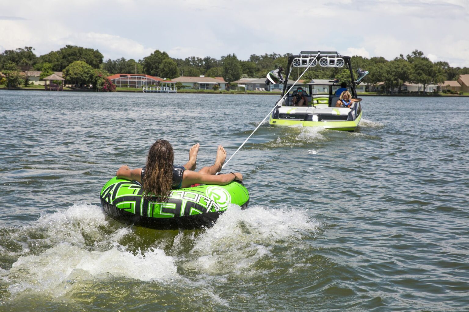 License-free image - A person with long hair rides an inflatable tube being towed by a boat on a lake. The green and black tube skims across the water, creating a splash. The boat ahead, which is white with green accents, is driven by another person. Houses and trees line the shore in the background. Tubing behind a wakeboard boat on Clear Lake in Orlando, Florida.