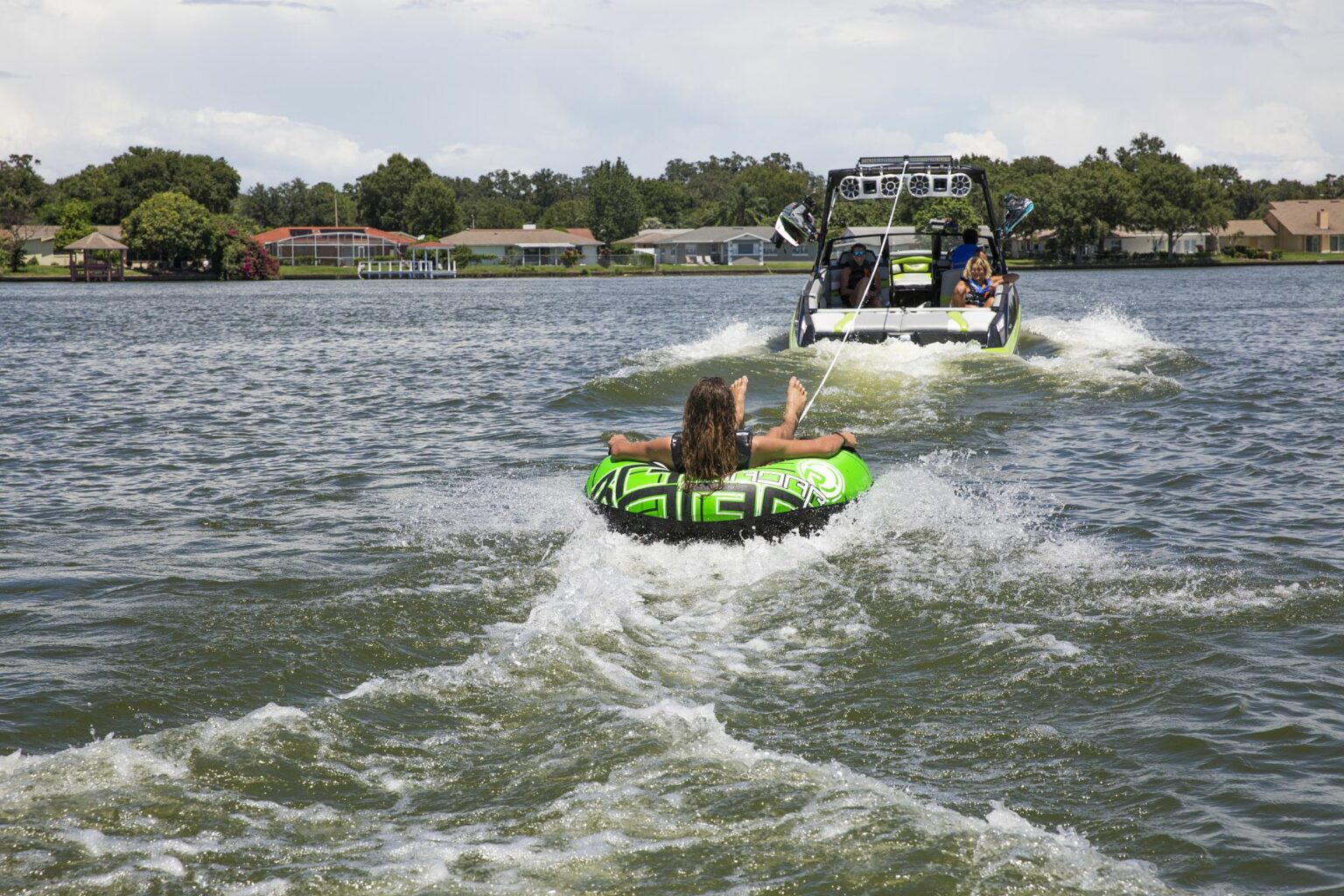 License-free image - A person rides on a green and black inflatable tube being towed behind a speedboat across a lake. The speedboat has several passengers, and houses are visible along the shoreline in the background, under a partly cloudy sky.