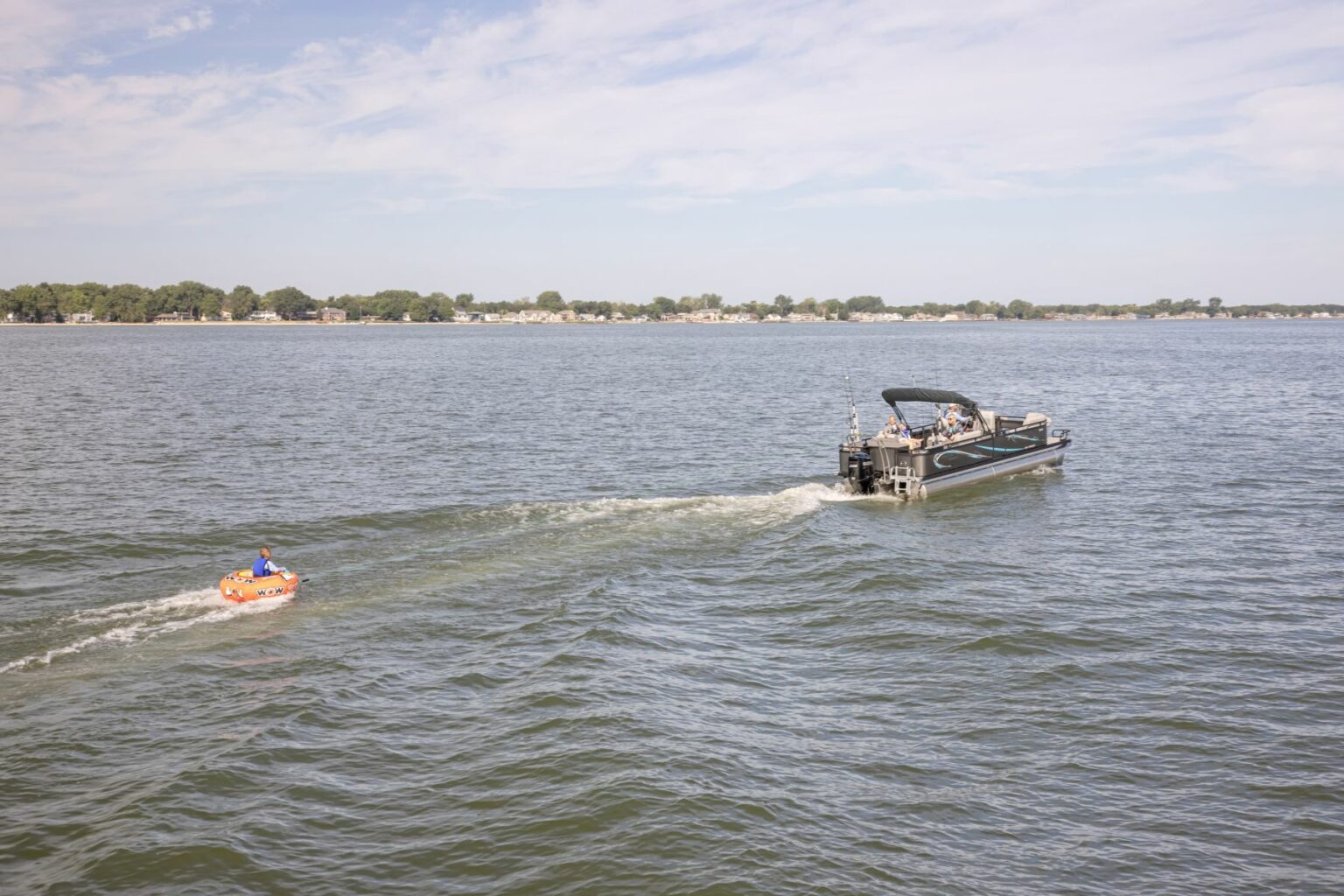 License-free image - A person on an orange inflatable tube is being towed by a black motorboat in a large body of water. The boat creates a wake as it moves forward. The horizon shows a distant shoreline with trees and buildings beneath a partly cloudy sky.