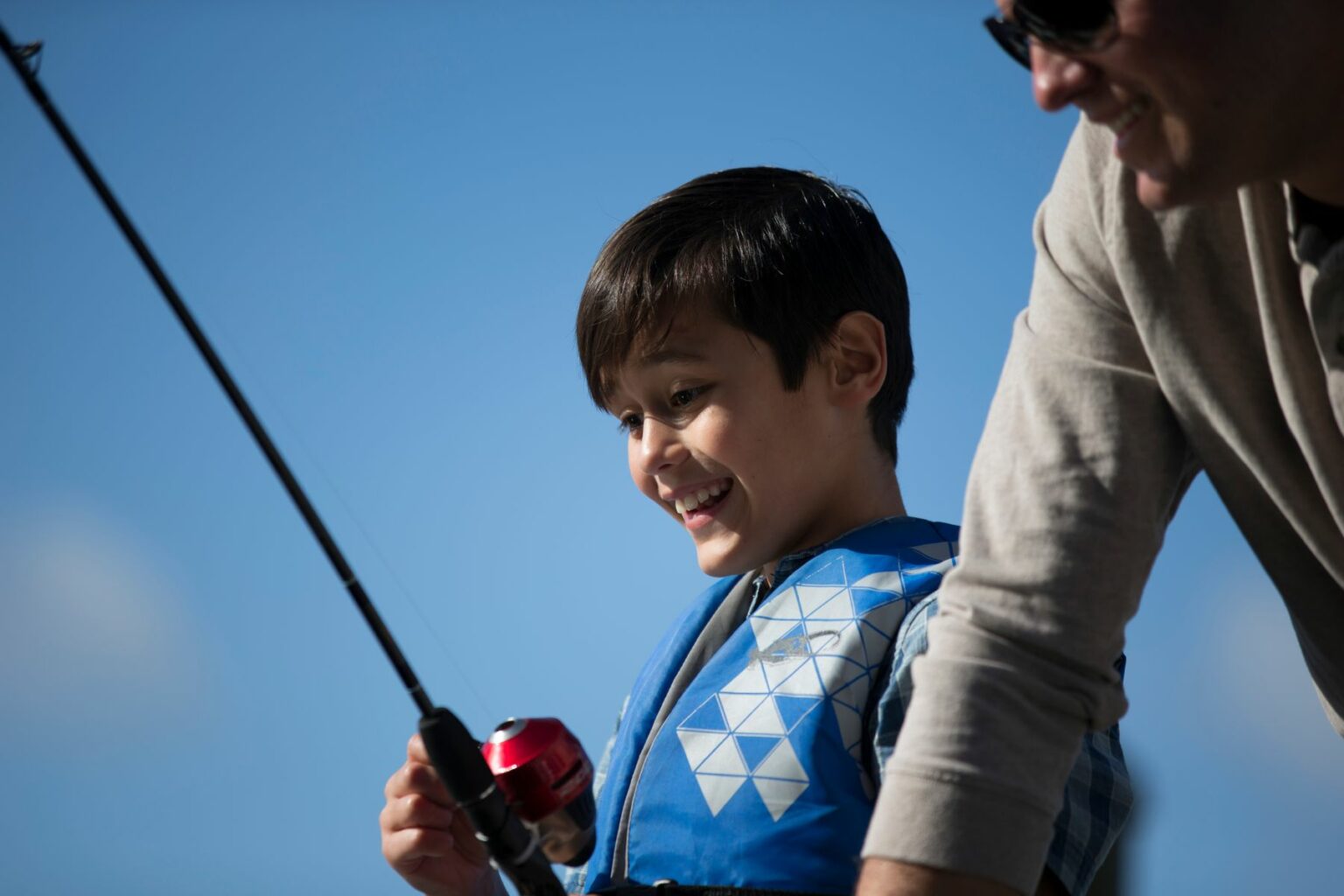 License-free image A smiling child wearing a blue life jacket holds a fishing rod, standing next to an adult in a beige sweater who is partly visible. The bright blue sky serves as the background, and both appear to be enjoying a day of fishing.