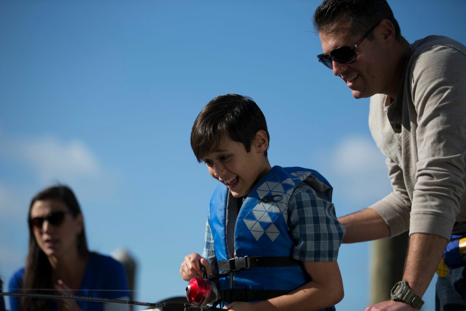 Royalty-free image A young boy wearing a blue life jacket and holding a fishing rod smiles excitedly. An adult man in sunglasses stands beside him, guiding him with his hand on the boy&#039;s shoulder. Another adult woman blurred in the background looks on. They are outdoors on a sunny day.