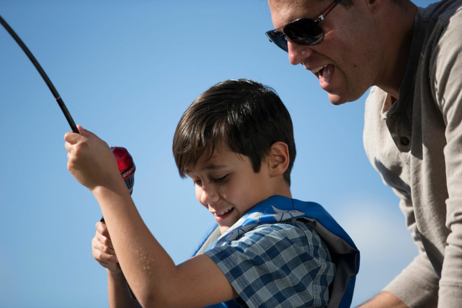 License-free image A man wearing sunglasses and a young boy in a blue life jacket are fishing together. The boy is smiling and holding a fishing rod, while the man enthusiastically guides him. The sky is clear and blue in the background.