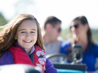 License-free image A young girl with long brown hair, wearing a red life jacket, smiles brightly at the camera. In the blurred background, two adults, a man and a woman, appear to be enjoying the day with her. The scene suggests a boat ride or outdoor activity.