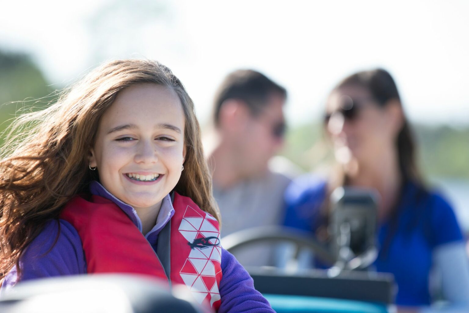 License-free image A young girl with long brown hair, wearing a red life jacket, smiles brightly at the camera. In the blurred background, two adults, a man and a woman, appear to be enjoying the day with her. The scene suggests a boat ride or outdoor activity.