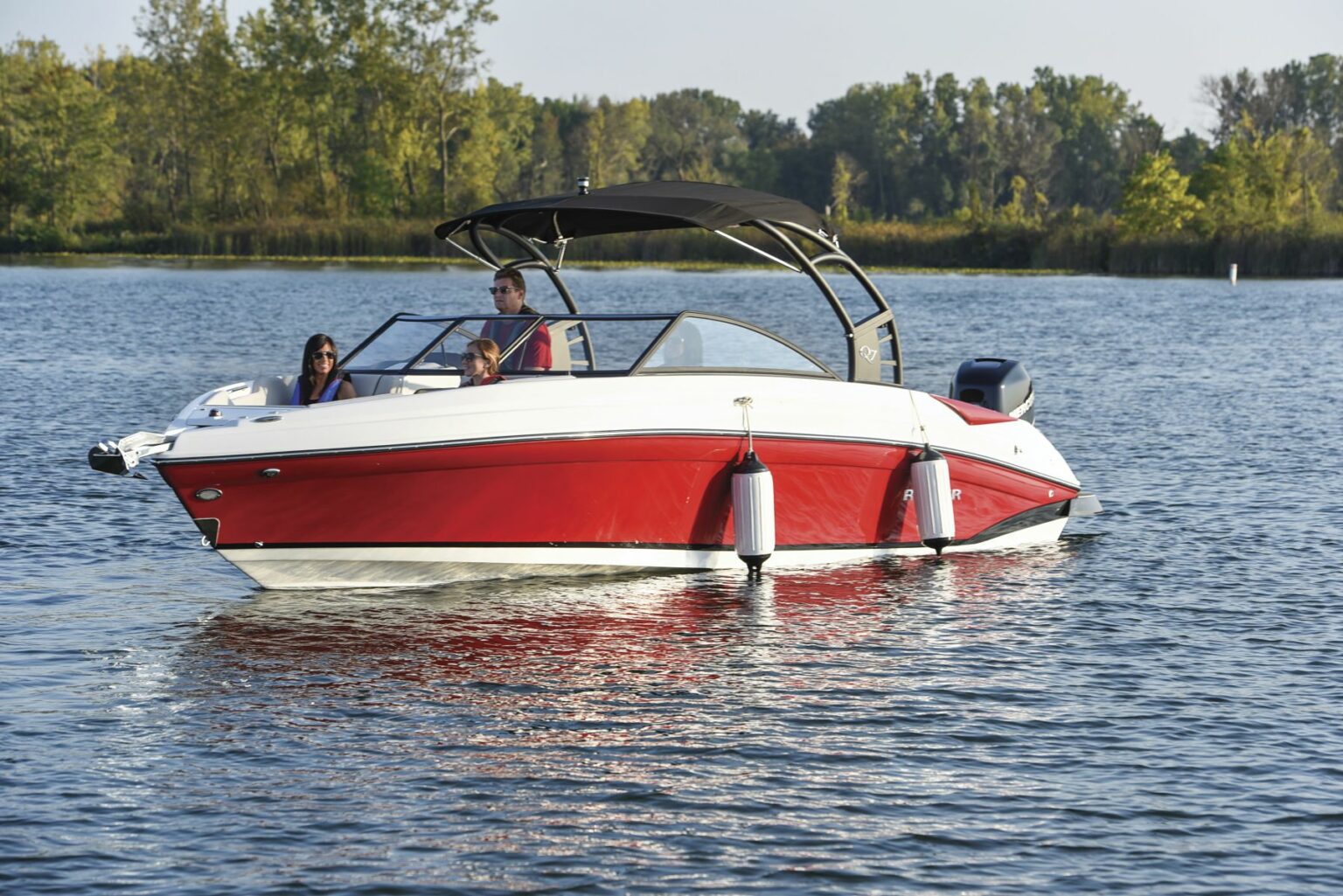 License-free image - Three people are on a sleek, red and white motorboat with a black canopy, floating on a calm lake surrounded by green trees. Two white fenders are hanging on the side of the boat to prevent damage. The sky is clear and blue. Preparing to dock a bay boat using fenders.