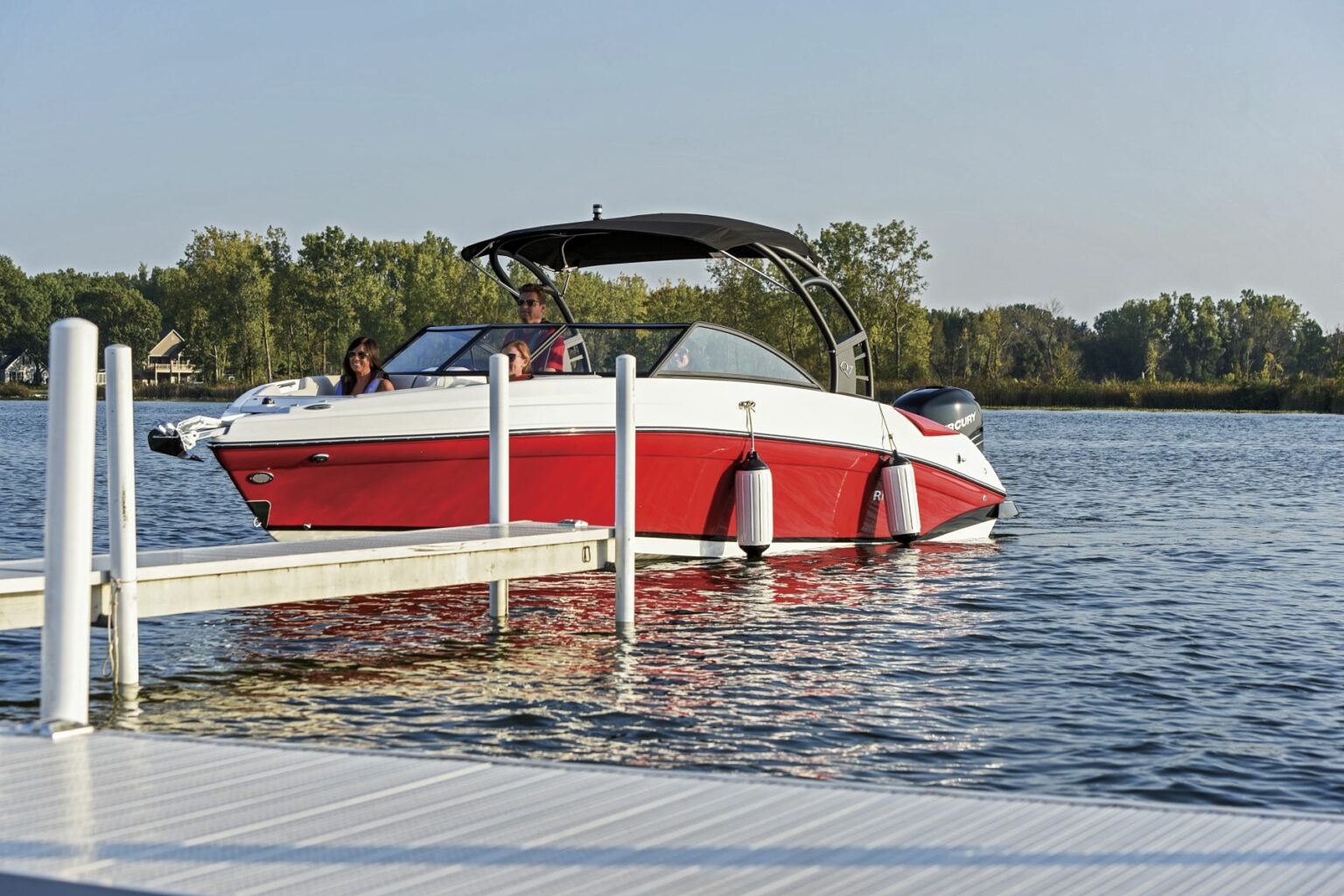 License-free image - A red and white boat with a black canopy floats near a dock on a calm lake. There are two people on board, one operating the boat and another seated nearby. Trees are visible in the background under a clear sky. Preparing to dock a bay boat using fenders.