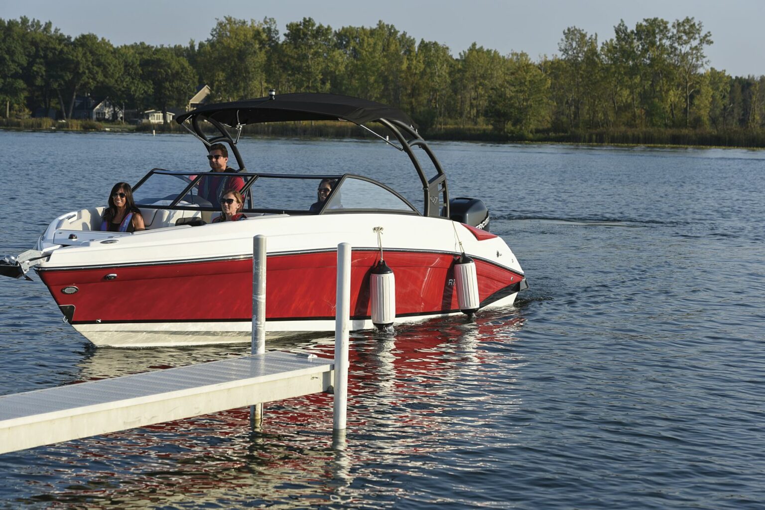 License-free image A red and white motorboat with a black canopy is seen on the water near a small dock with white bumpers. Three people are sitting on the boat, enjoying a sunny day, with lush green trees visible in the background.