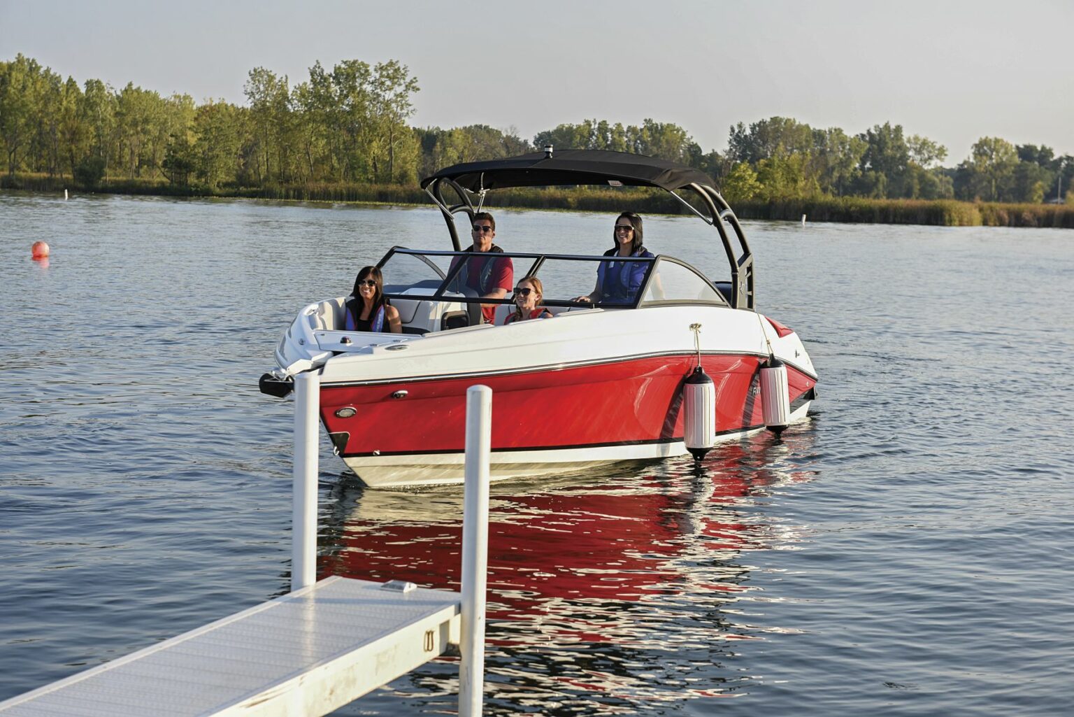 License-free image - A red and white motorboat with a black canopy is on a lake near a dock. Four people are on the boat, wearing casual clothes and life jackets. Trees and greenery are visible in the background under a clear sky. Preparing to dock a bay boat using fenders.