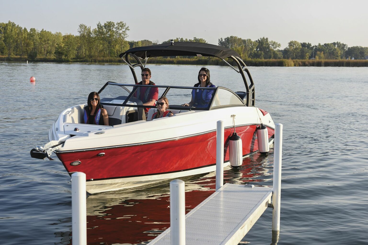 License-free image - A group of five people are sitting on a red and white motorboat, which is docked beside a pier. The water is calm, and trees are visible in the background on a sunny day. The individuals are wearing life jackets and appear to be enjoying their time on the boat. Preparing to dock a bay boat using fenders.