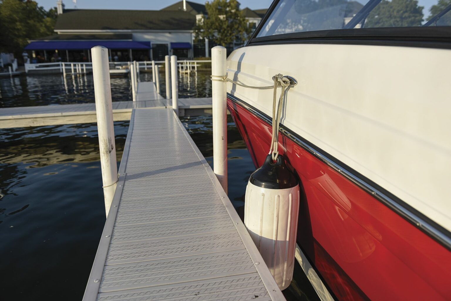 License-free image - A red and white boat is docked at a marina. The boat is secured to the dock with a rope, and there is a white fender hanging on its side. The dock is made of metal, leading out towards the water, with reflections of trees and buildings in the background. Preparing to dock a bay boat using fenders.