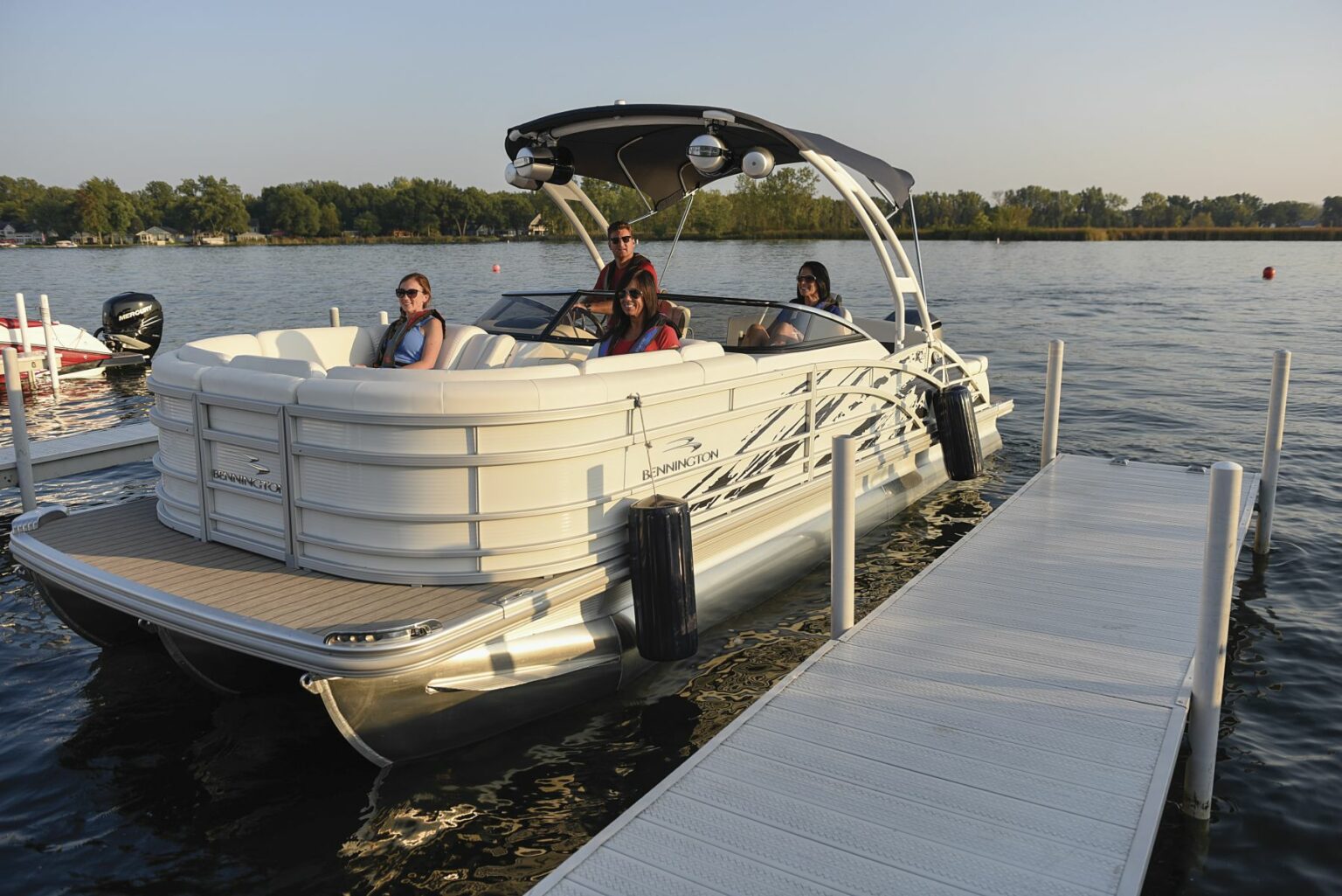 License-free image - A group of people sit on a white Bennington pontoon boat docked on a calm lake. The boat is near a white floating dock, surrounded by still water reflecting the clear sky. Trees and greenery are visible in the background. Preparing to dock a pontoon boat using fenders.