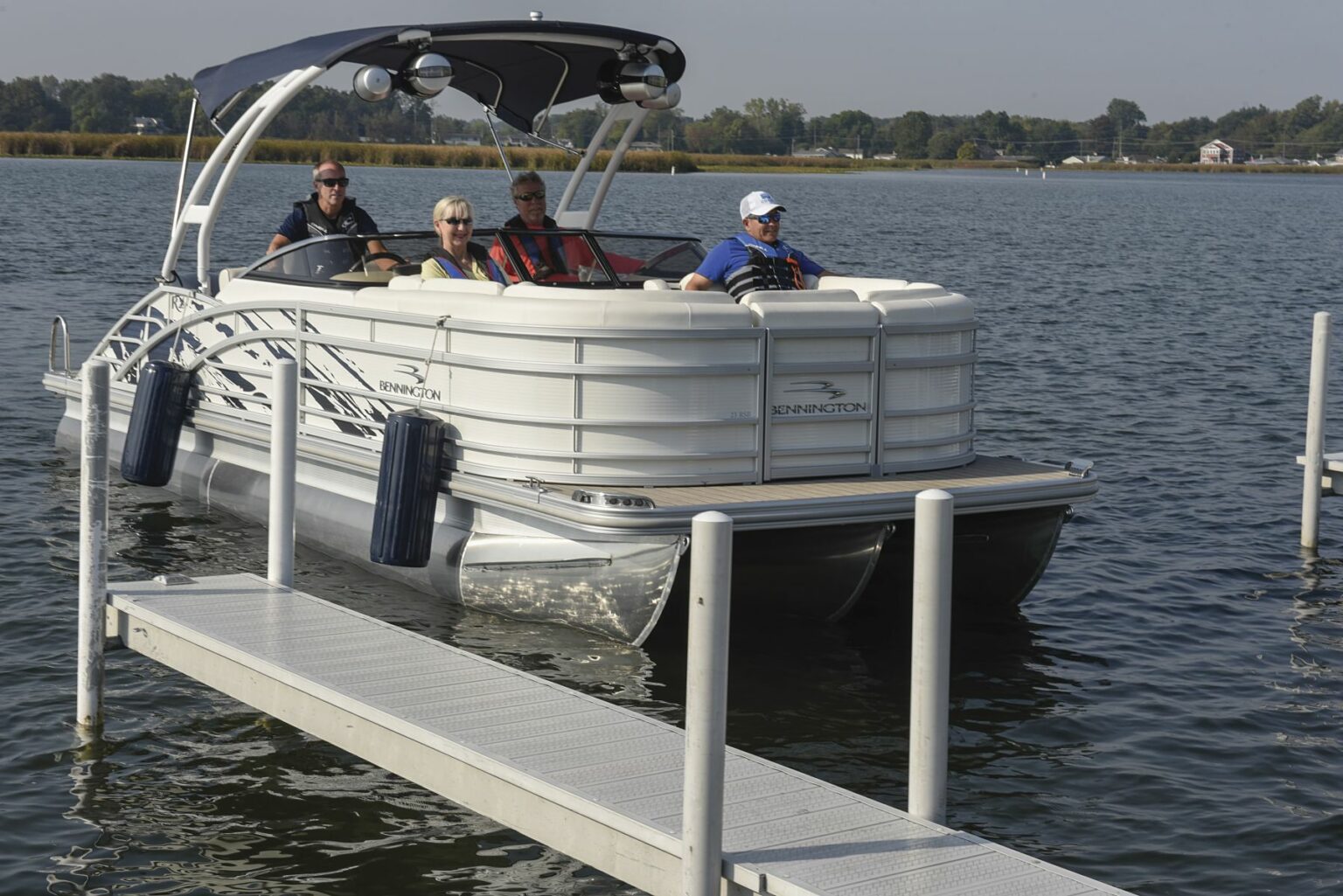 License-free image - A pontoon boat with four people is approaching a dock on a calm lake. The boat has a canopy, and the passengers, dressed in casual clothes and life jackets, are seated comfortably. The surrounding area has trees and distant houses along the shore. Preparing to dock a pontoon boat using fenders.