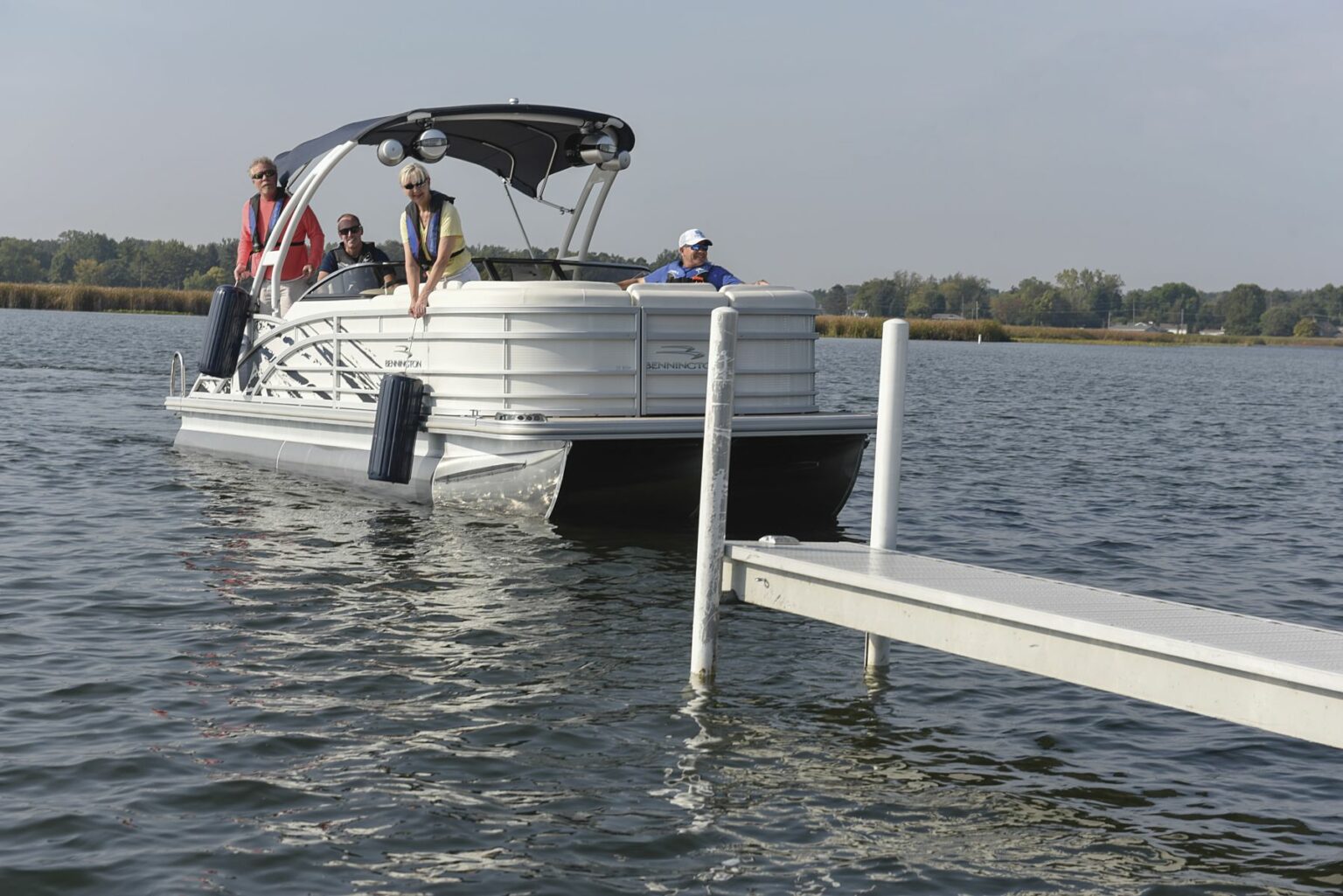 License-free image - A pontoon boat with three people on board is approaching a dock on a calm lake. The boat has a canopy and safety equipment visible. The passengers, wearing casual clothes, appear to be preparing to dock. The background shows a shoreline with trees. Preparing to dock a pontoon boat using fenders.