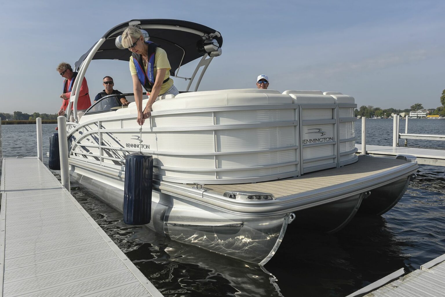 License-free image - A group of four people on a white pontoon boat docked at a pier. One person wearing a life jacket is leaning over the side of the boat, adjusting a fender to protect the boat from the dock. The scene is set against a calm body of water on a sunny day. Preparing to dock a pontoon boat using fenders.