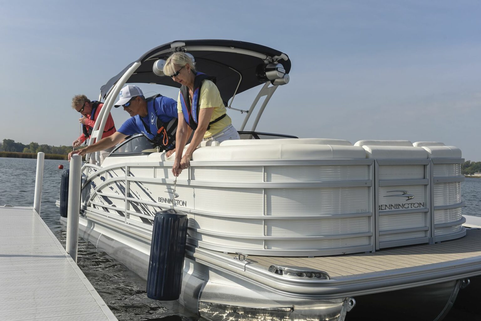 License-free image - Three individuals are docking a white Bennington pontoon boat. Two people on board, wearing life jackets, are securing the boat with ropes, while another person stands on the dock assisting. The scene is set on a calm body of water with a clear sky. Preparing to dock a pontoon boat using fenders.