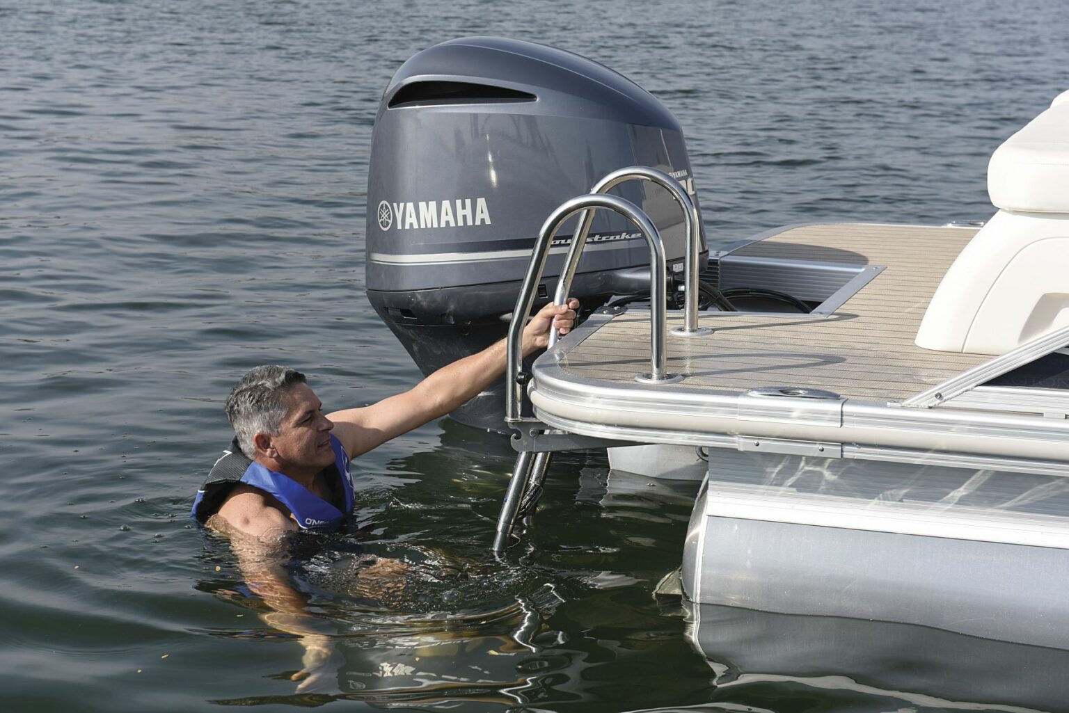 License-free image - A man in a blue life vest is in waist-deep water, holding onto a metal ladder attached to a boat. The boat has a Yamaha outboard motor and is floating on a calm body of water. The man appears to be about to climb onto the boat.