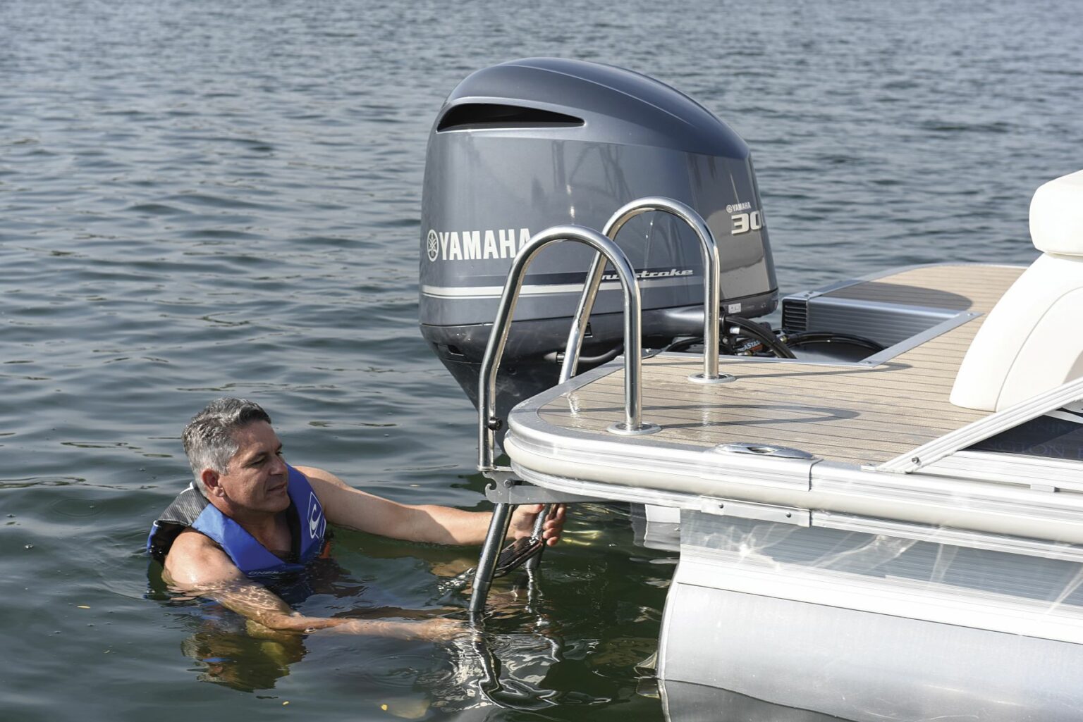 License-free image - A man wearing a blue life jacket is in the water near a boat, holding onto the ladder of the boat&#039;s engine platform. The boat has a Yamaha outboard motor, and the man appears to be getting ready to climb back onto the boat. The water is calm and clear.