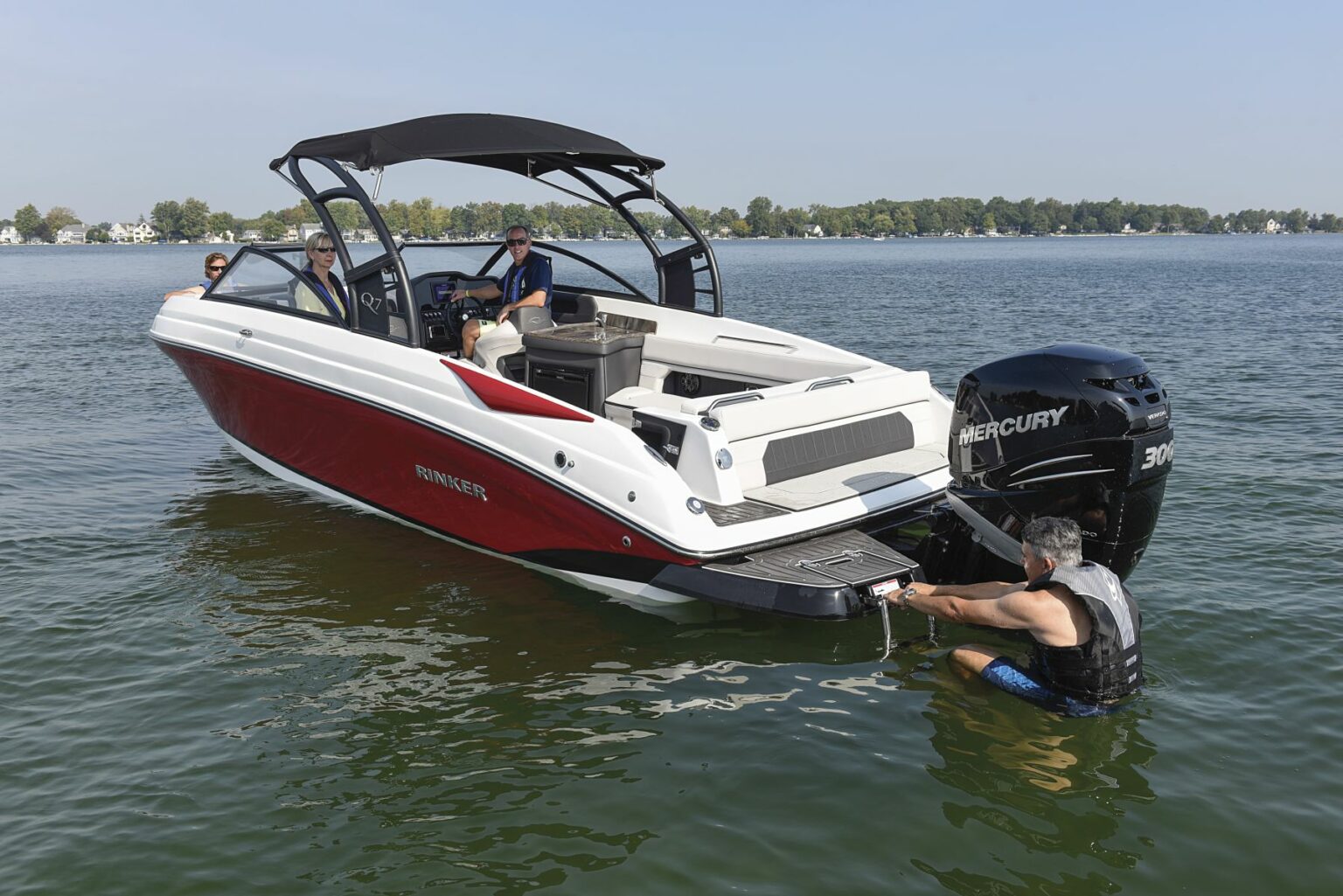 License-free image - A red and white motorboat with four people onboard is seen in a calm body of water. One person is climbing a ladder at the back of the boat wearing a USCG approved life vest. The boat has &quot;Rinker&quot; branding and is powered by a Mercury 300 outboard engine. Trees and houses are visible in the background.