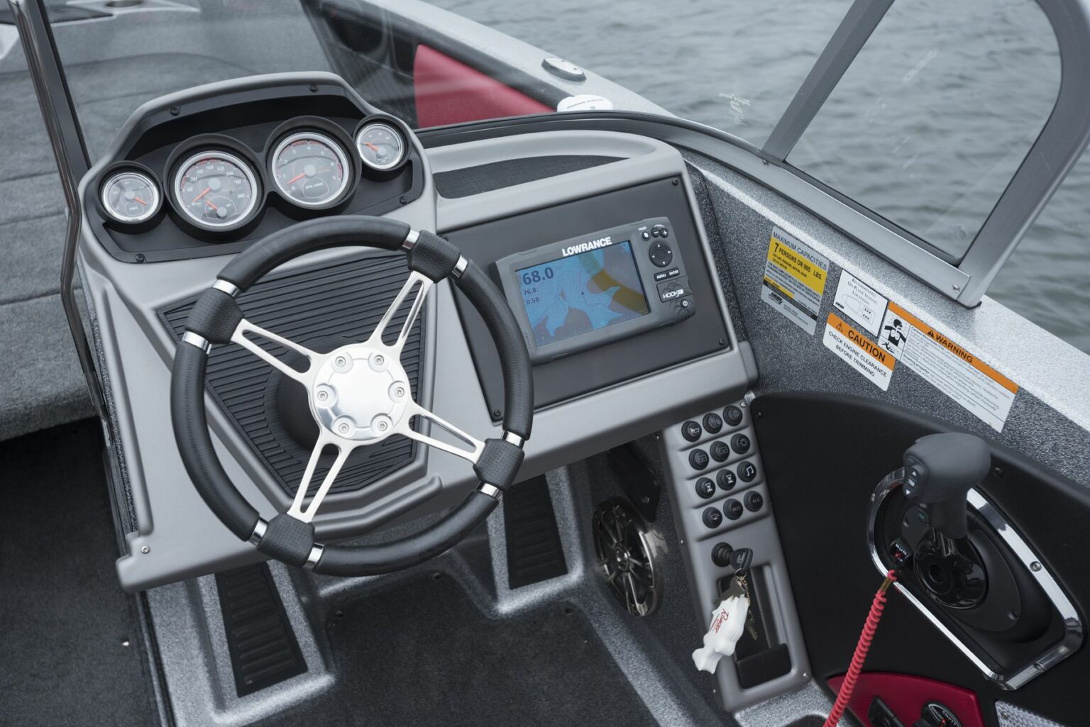 License-free image - A boat&#039;s helm with a steering wheel, multiple gauges, and a Lowrance fish finder on the console displaying a waterway map. The throttle control and a red safety kill switch cord are also visible on the right side of the console. A capacity plate and certificate of compliance.