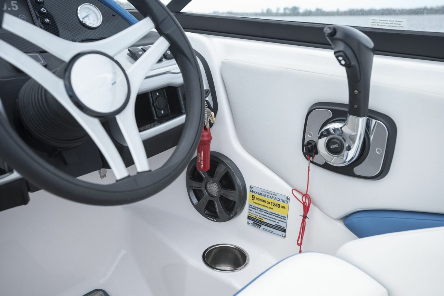 License-free image - Close-up of the driver&#039;s station on a boat, featuring a white steering wheel, throttle control with a black and white handle, a speaker, and a safety lanyard with a red clip. Various warning and instruction stickers are visible near the control panel. A capacity plate and certificate of compliance.