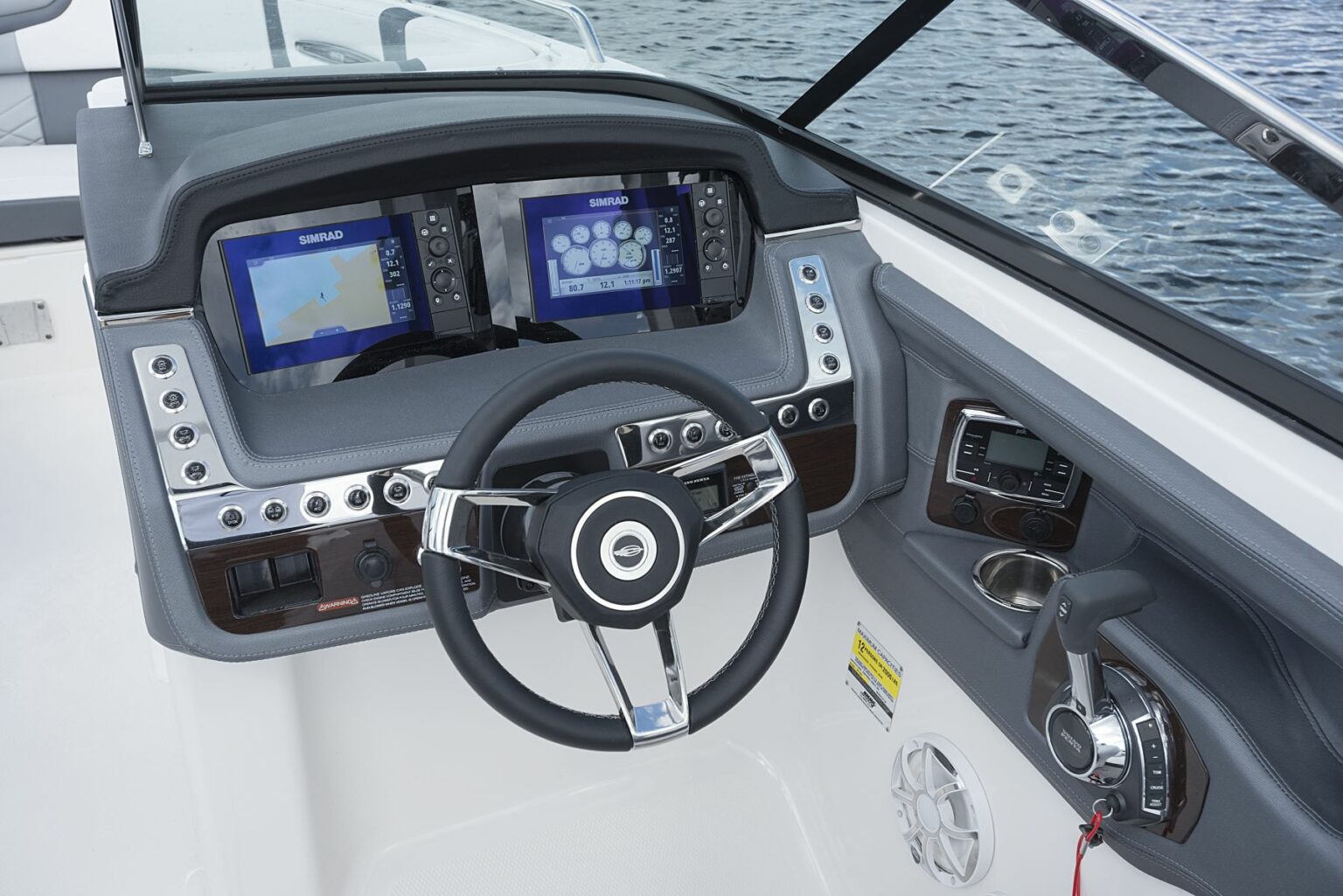 License-free image - The helm of a modern motorboat featuring a steering wheel, dual Simrad display screens, and various navigational and control buttons. The throttle control and a speaker are on the right side of the console. Water is visible in the background. A capacity plate and certificate of compliance.