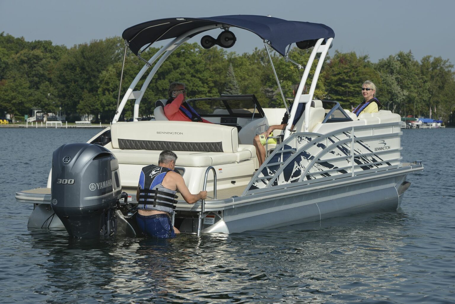 License-free image - A man in a life jacket climbs the ladder of a pontoon boat named Bennington with a Yamaha 300 engine. Three people are sitting onboard, wearing casual summer clothing and enjoying the water. Trees and other boats are visible in the background.