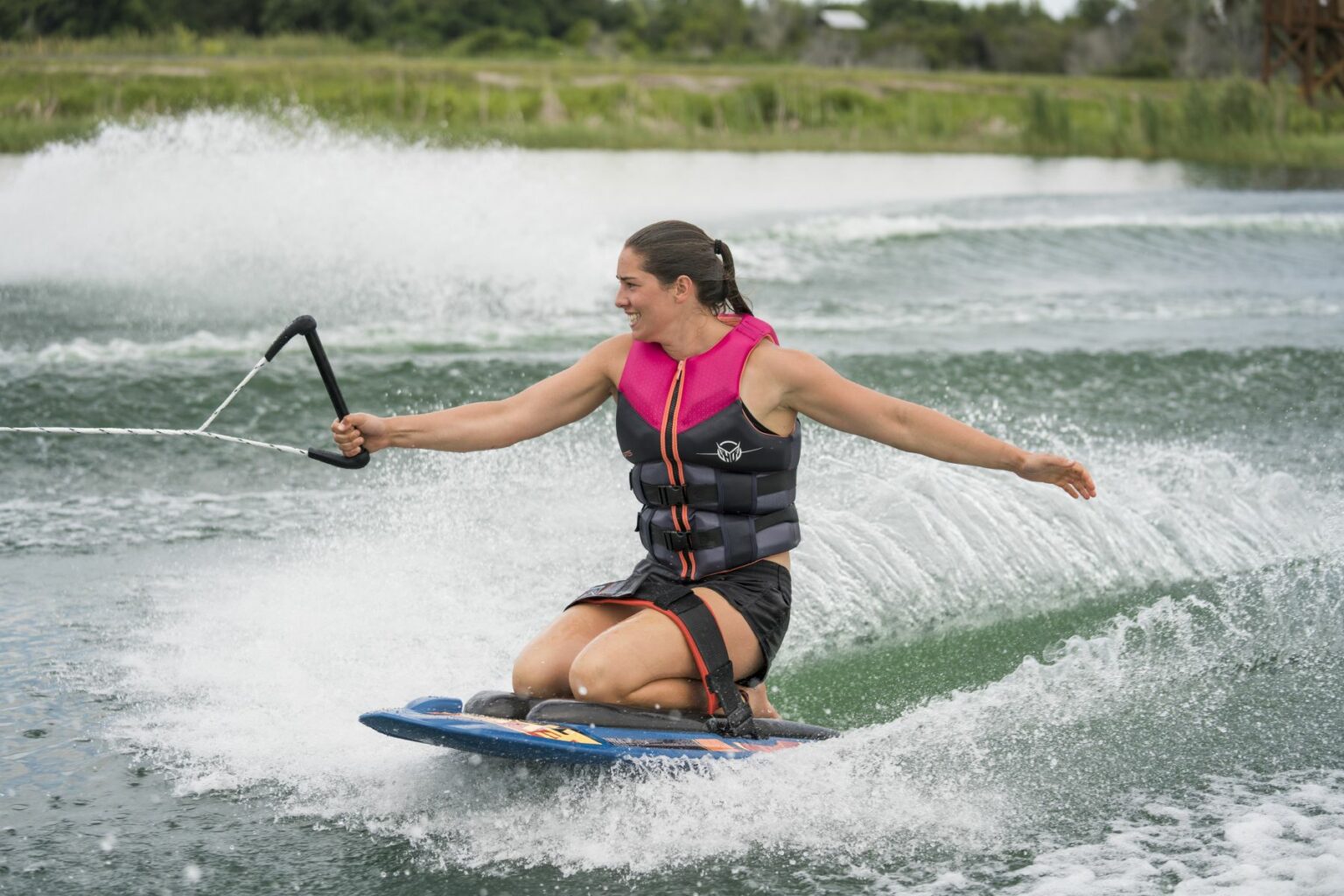 License-free image - A woman is kneeboarding on a body of water, holding onto a tow rope with one hand and balancing with the other. They are wearing a black and pink life vest, and there are splashes of water around them. Lush greenery is visible in the background.. Shoot in Orlando and Miami Florida