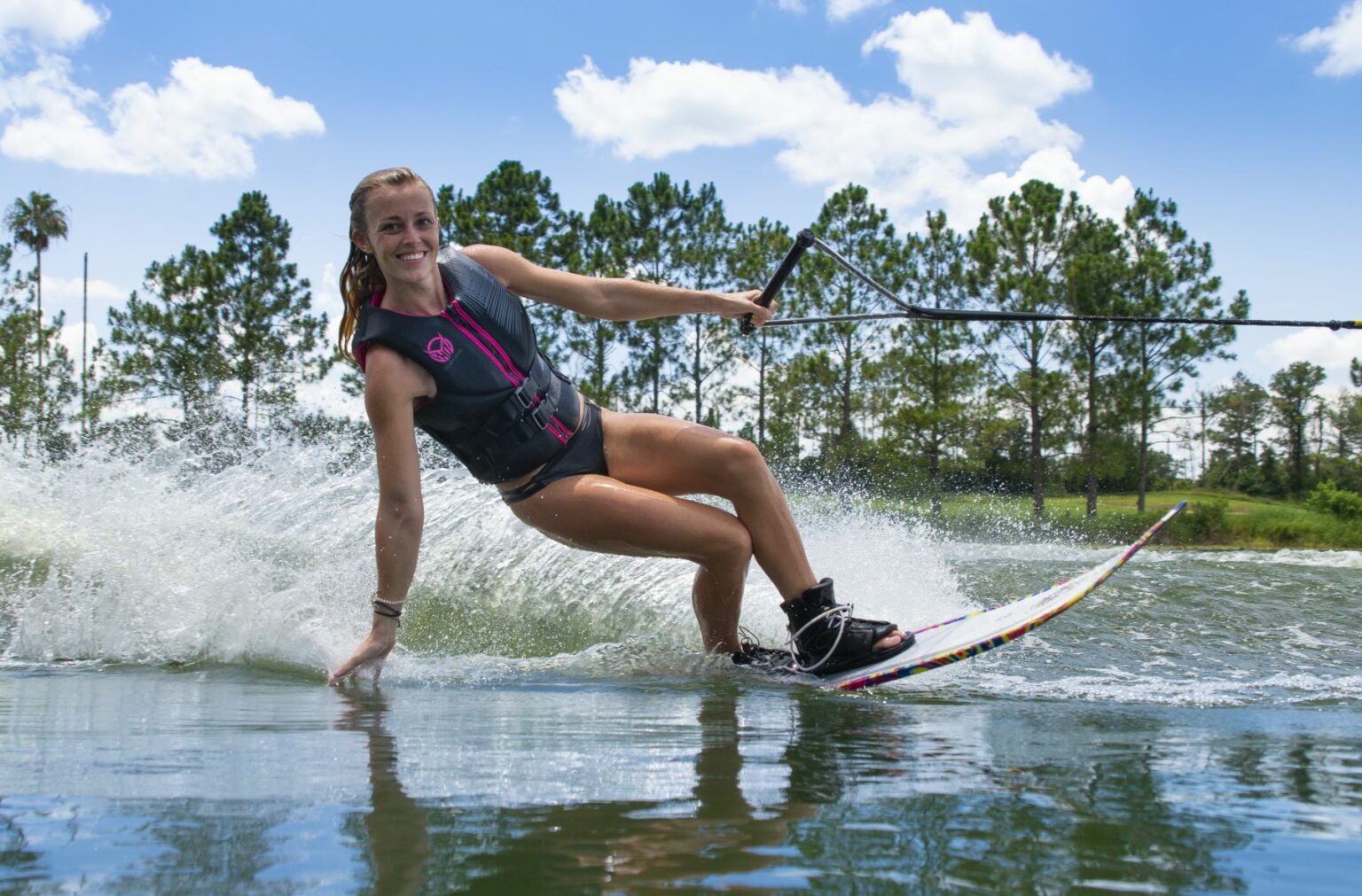 License-free image - A woman wearing a life jacket skillfully slaloms on a waterski across a lake, holding onto a rope handle. They smile at the camera, creating a splash behind them. The background features green trees and a blue sky with scattered white clouds.