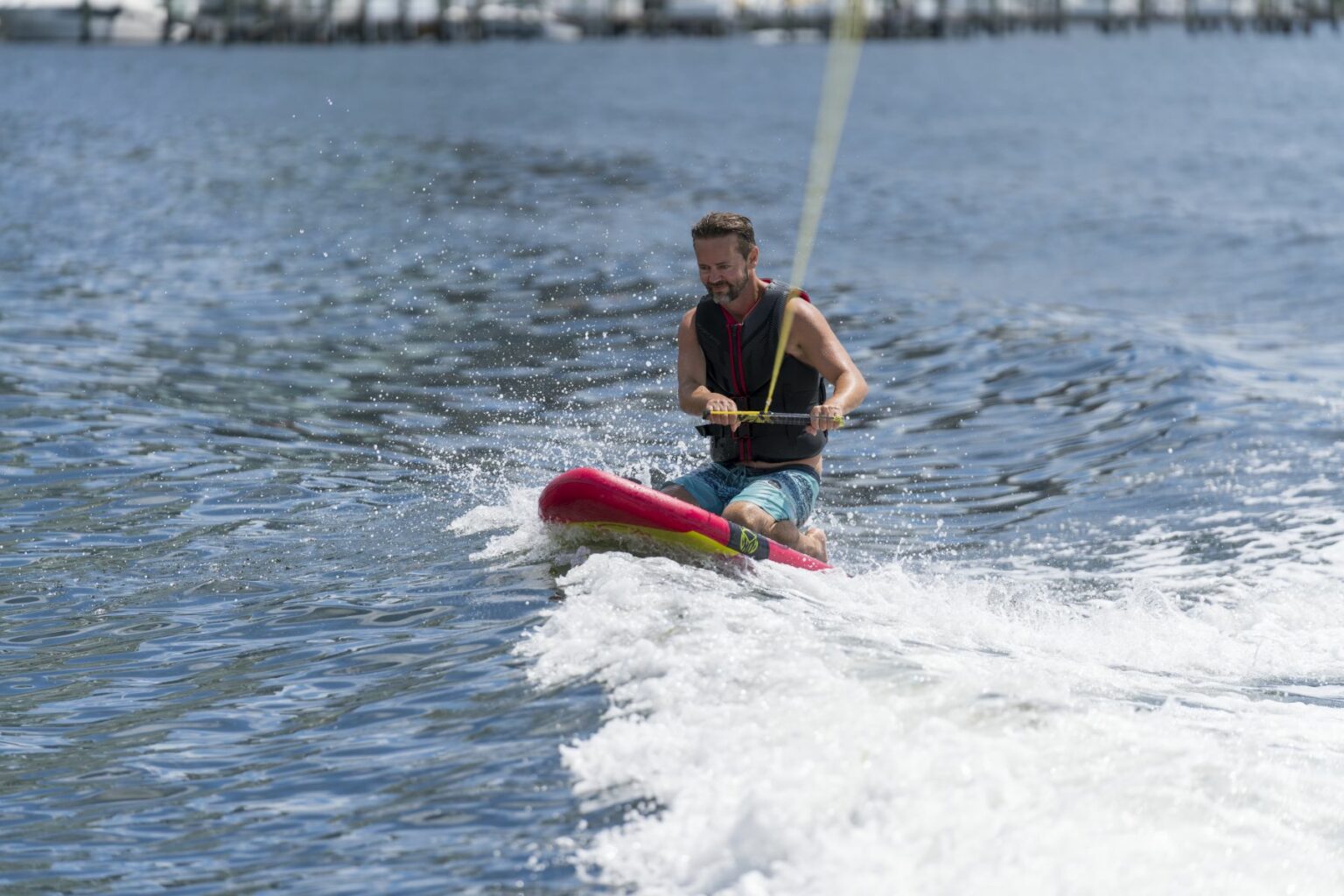 License-free image - A man wearing a life vest rides a kneeboard on the water, gripping a tow rope. The water is splashing around him as he navigates the waves, with the background showing a distant shoreline and docked boats. Shoot in Orlando and Miami Florida watersports, waterskiing