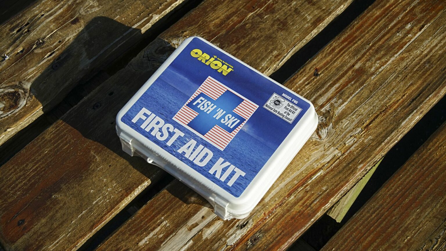 License-free image - A white first aid kit with &quot;First Aid Kit&quot; printed on the front, featuring a red cross and a blue background with a fish motif. The kit is branded &quot;Orion&quot; and labeled &quot;Fish &#039;N Ski.&quot; The kit is placed on a weathered wooden surface. A first aid kit, essential equipment on vessels of any size.