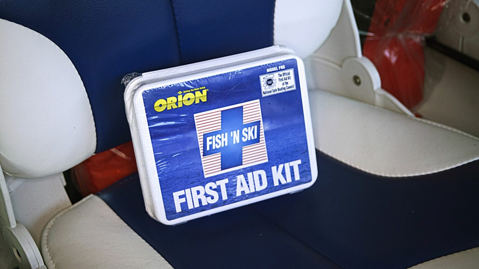 License-free image - A blue and white first aid kit labeled &quot;Fish &#039;n&#039; Ski&quot; and &quot;Orion&quot; is placed on a blue and white cushioned boat seat. The kit is designed for boating or fishing emergencies, providing essential medical supplies. A first aid kit, essential equipment on vessels of any size.