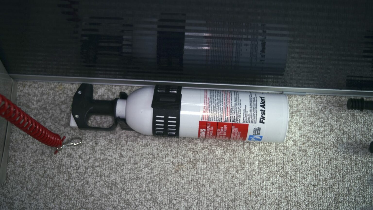 Royalty-free image - A white fire extinguisher lies on a beige carpeted floor beside a corrugated black metal panel. The extinguisher features a red and black label with safety instructions, and a black nozzle with a hose attached, leading to a red cord. A mounted fire extinguisher, easily accessible in the event of an emergency.