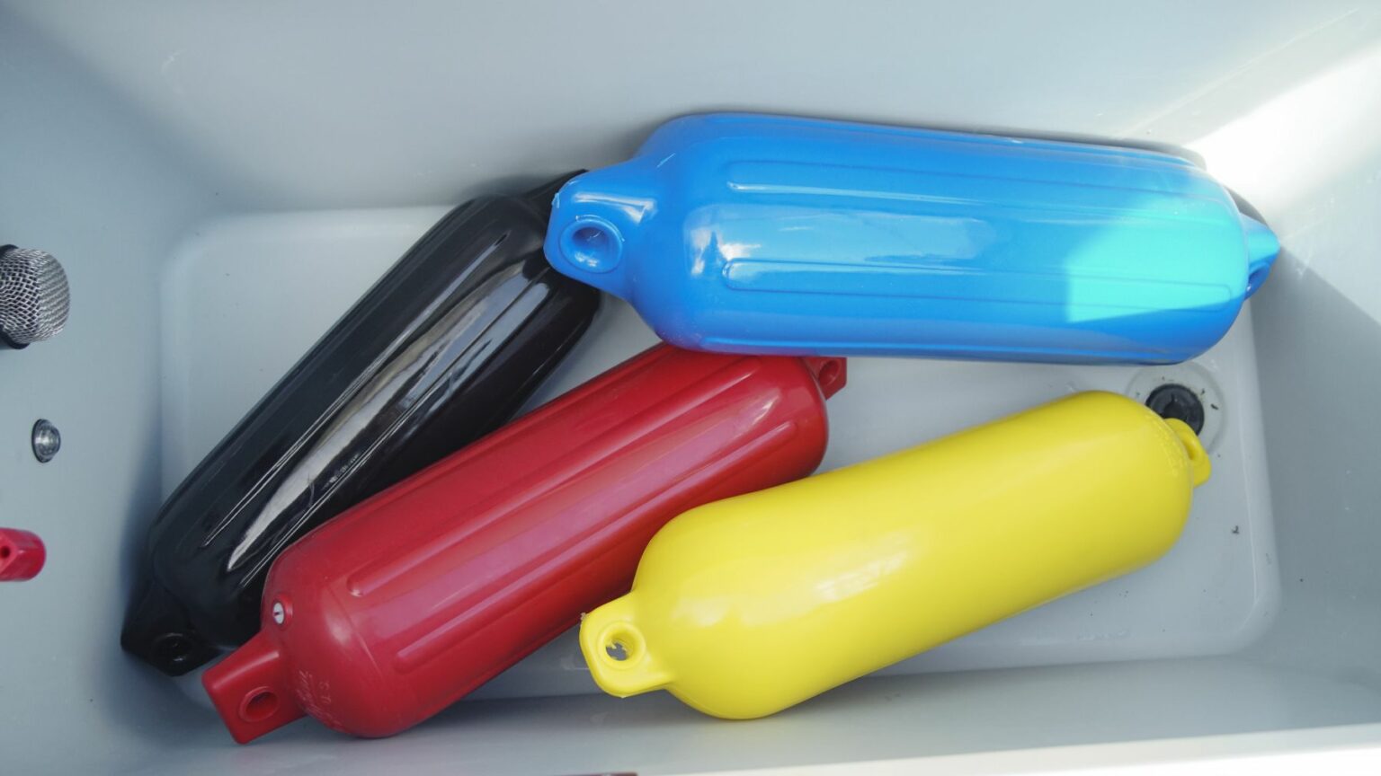 License-free image Four colorful fenders in black, red, blue, and yellow are placed inside a white container. The fenders are cylindrical with rounded ends and have a small hole at one end for attachment. Fenders stowed aboard a boat.