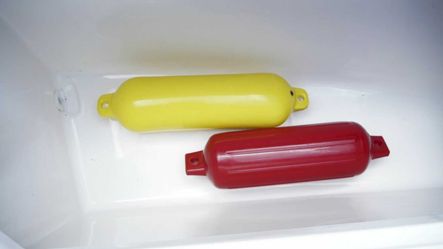 License-free image - A yellow and a red cylindrical buoy like boat fender are placed inside a white container. Both buoys have small holes for attachment on their ends. The container has smooth, shiny surfaces. Fenders stowed aboard a boat.