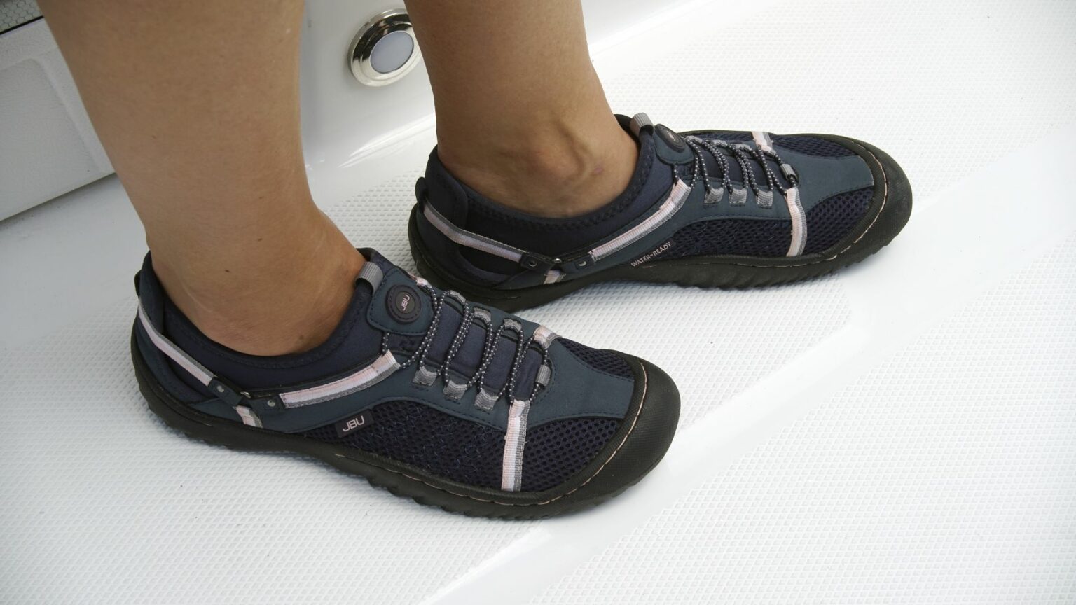 License-free image - Feet wearing dark blue athletic shoes with pink stripes and black soles, standing on a shiny white surface. The shoes feature mesh fabric and elastic laces with a fastener. Boat shoes are essential gear on any sized vessel.