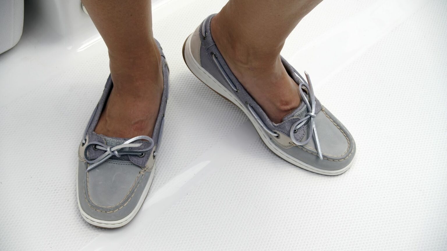 License-free image - Close-up of a person&#039;s lower legs and feet wearing gray boat shoes with white laces and stitching, standing on a white textured surface. Boat shoes are essential gear on any sized vessel.