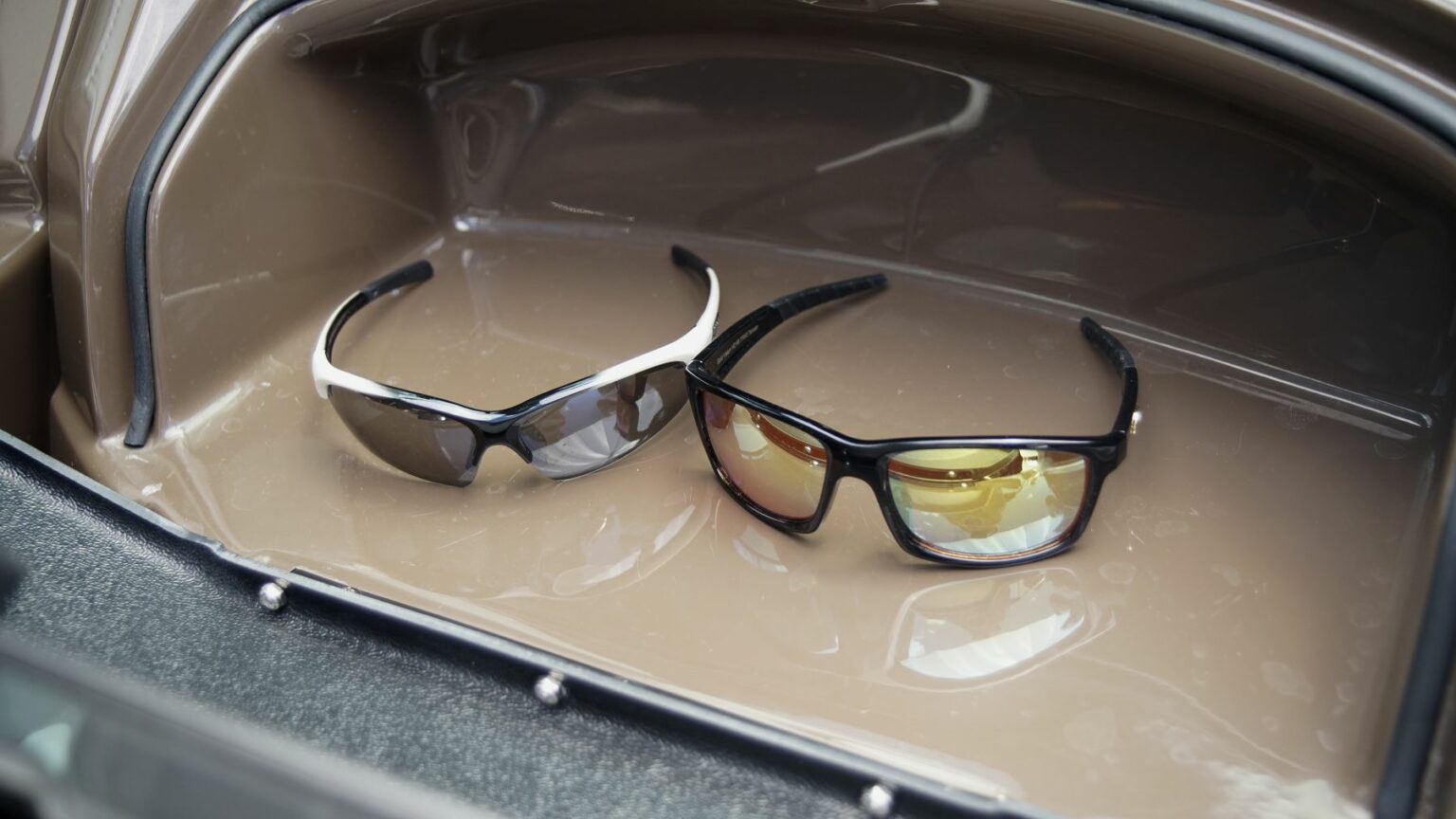 License-free image -  A pair of sunglasses inside a compartment. The left pair has white frames and dark lenses, while the right pair has black frames with yellow-tinted lenses. The surface inside the compartment appears to be shiny and smooth. Sunglasses are essential gear on any sized vessel.