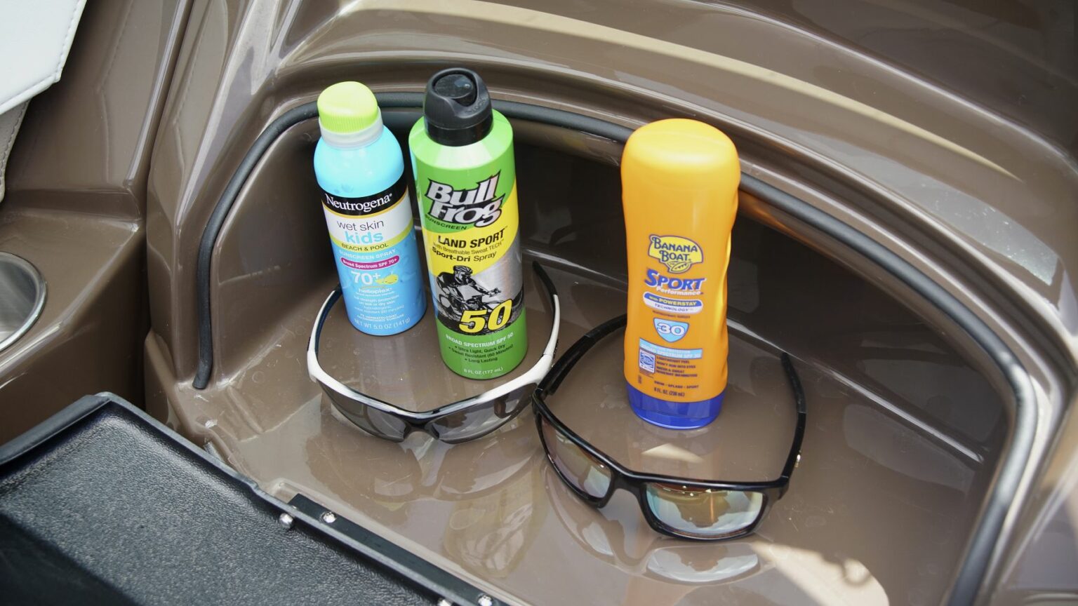 License-free image - Three bottles of sunscreen and two pairs of sunglasses sit in a brown compartment. The sunscreens include Banagard Kids SPF 70, Bull Frog Land Sport SPF 50, and Banana Boat Sport SPF 30. The sunglasses have black frames and are placed next to each other. Sunglasses and sunscreen is essential equipment on any sized vessel.