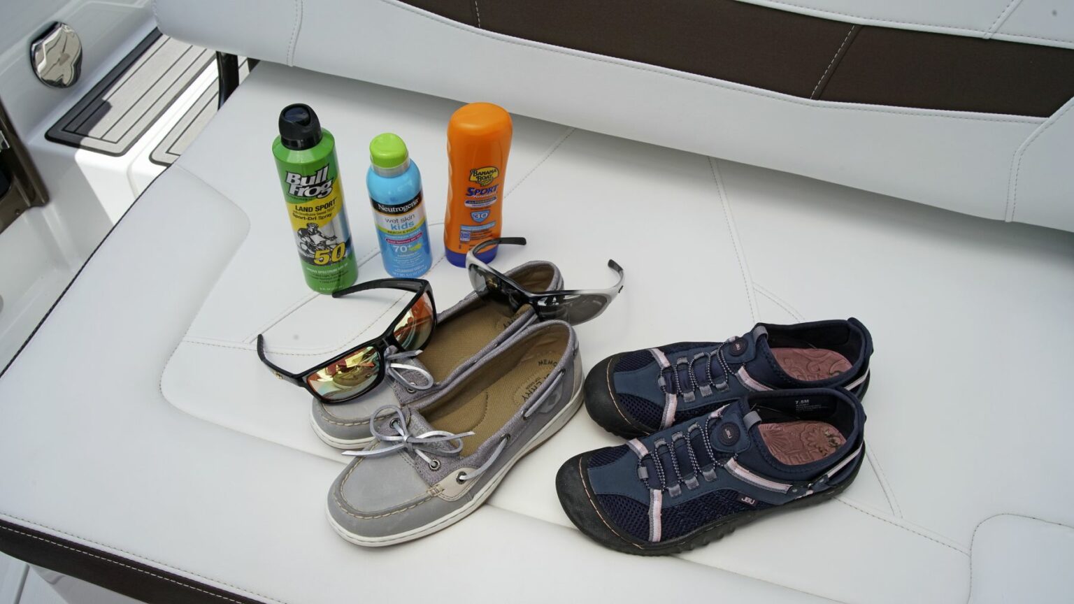 License-free image - A pair of sunglasses and eyeglasses, casual gray boat shoes, and navy blue sports shoes are placed on a white cushioned seat. Next to them are three bottles: BugStop insect repellent, Banana Boat sunscreen, and an orange can of sunscreen spray. Sun gear is essential equipment on any sized vessel.