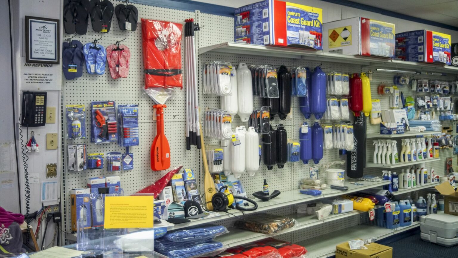 License-free image -  A store shelf displays various boating supplies, including paddles, life jackets, fenders, ropes, and cleaning products. The items are neatly arranged on white pegboard shelves with some additional products on an adjacent counter. Safety equipment on sale in a retail outlet.