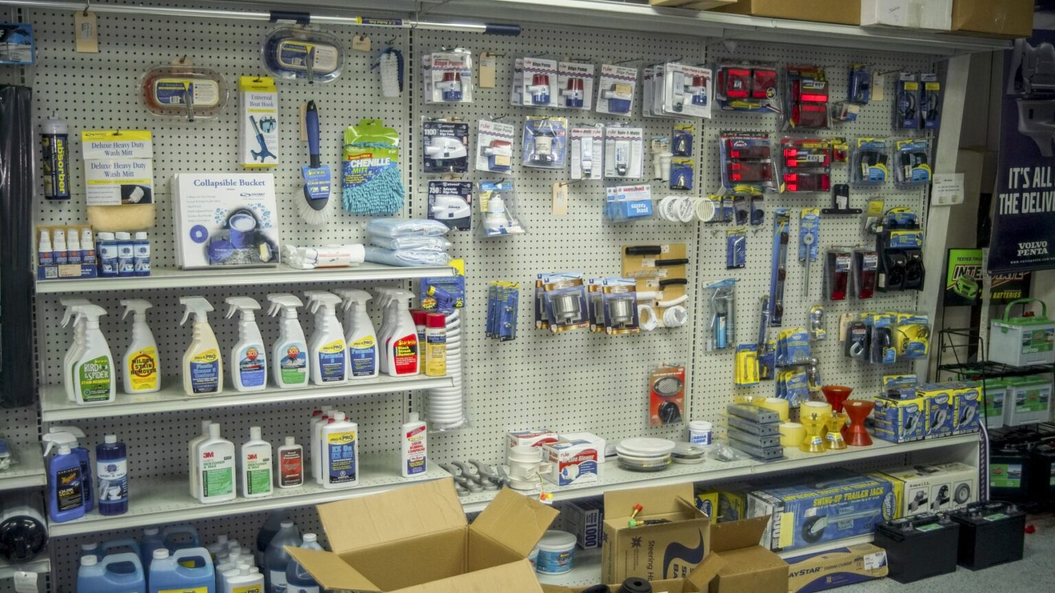 License-free image - An array of various household and hardware items displayed on shelves in a store. The items include cleaning sprays, light bulbs, electrical supplies, hand tools, batteries, and other miscellaneous utensils. Two open cardboard boxes are on the floor in the foreground. Safety equipment on sale in a retail outlet.