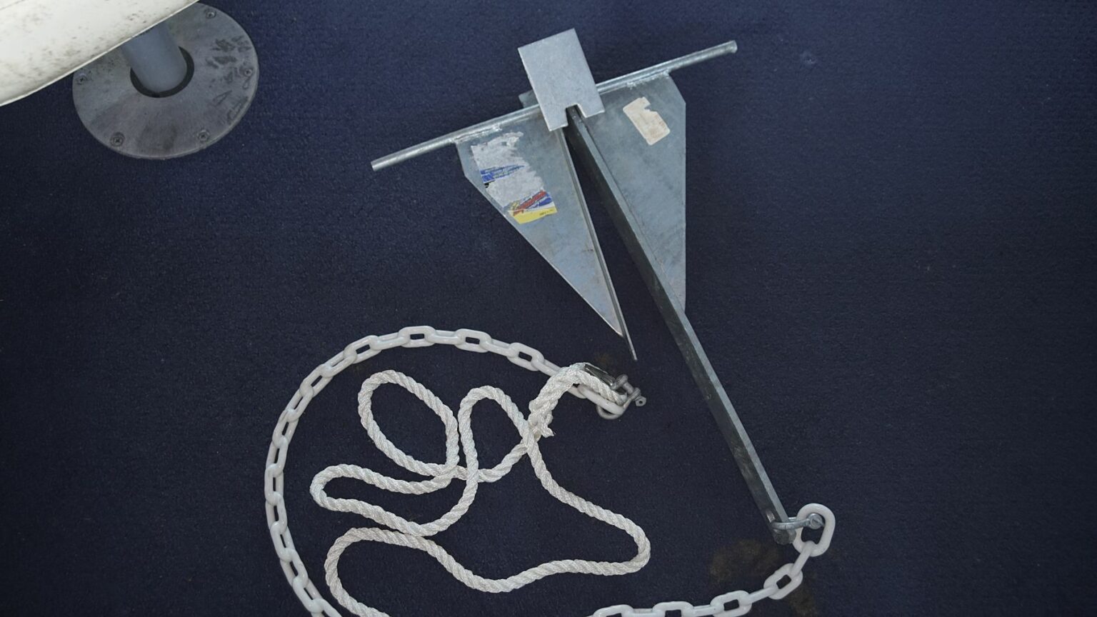 License-free image - A metal boat anchor with flukes and a stock is lying on a dark blue surface. A white rope and chain are attached to the anchor, coiled on the ground. Part of a metallic table base is visible in the upper left corner. A fluke, danforth or lightweight anchor.