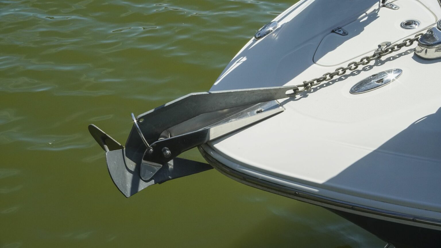 Royalty-free image - Close-up of the bow of a white boat with a metal anchor attached at the front. The anchor is connected by a chain and rests on a mount. The boat is on greenish water, and the sunlight is creating shadows on the boat&#039;s surface.