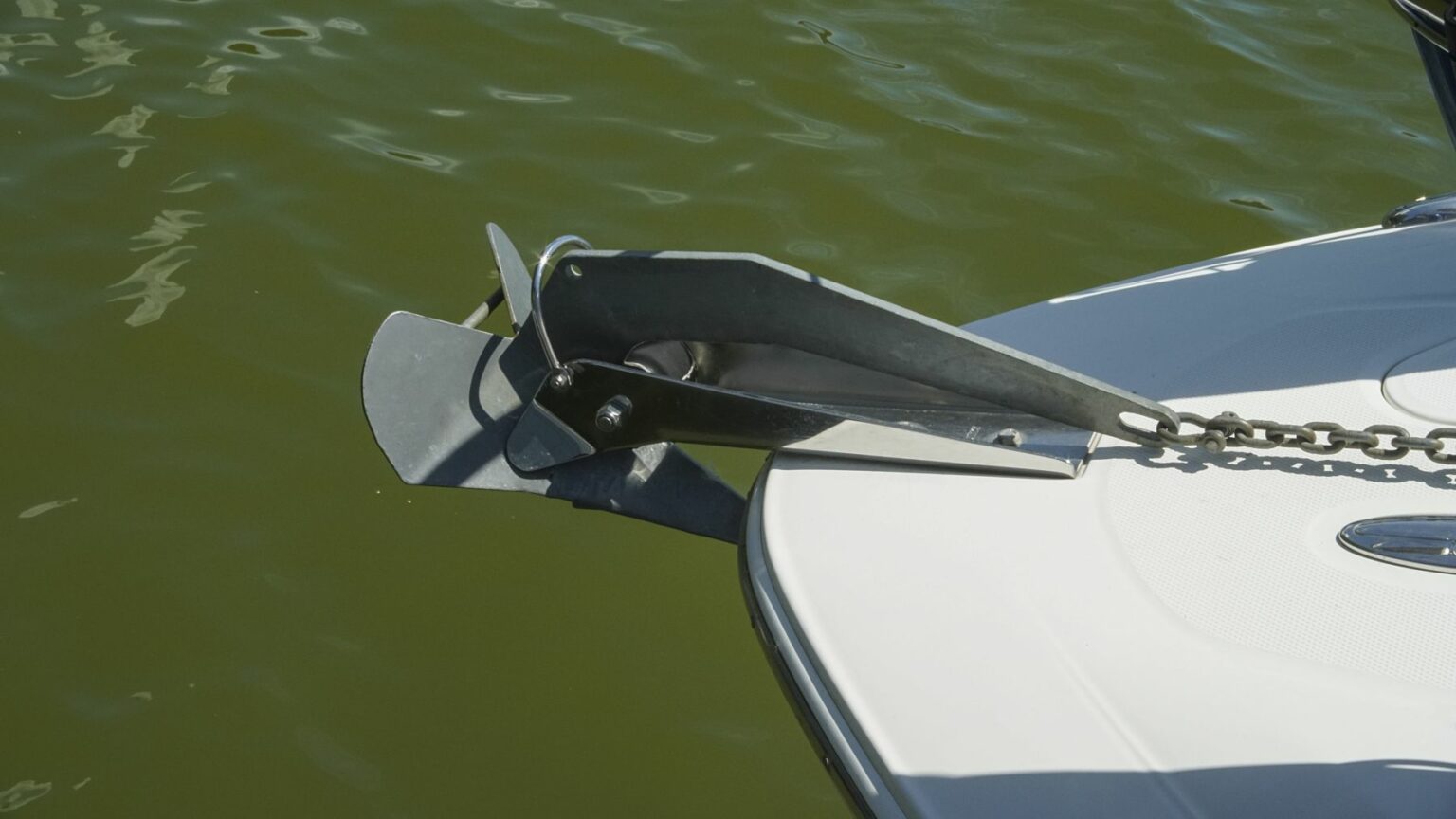 License-free image - A close-up of a boat&#039;s anchor secured to the bow, partially submerged in greenish water. The metal anchor is attached to a chain and is positioned at the edge of the boat&#039;s white surface. A plow, CQR, wing or delta anchor.