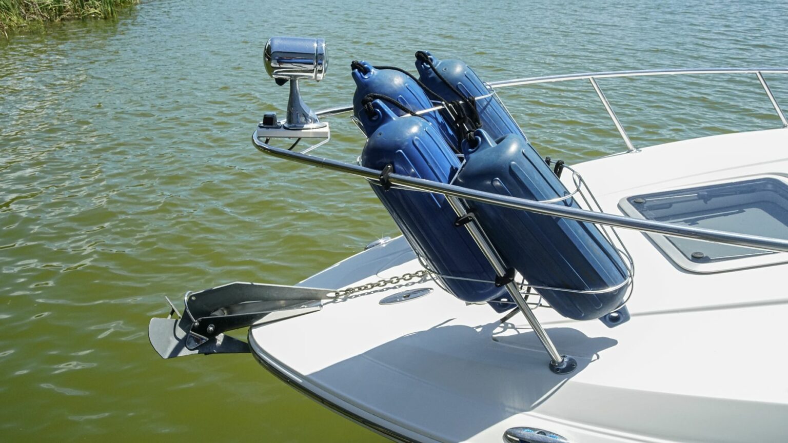 License-free image - View of the bow of a white boat with blue fenders attached to the rails. A stainless steel anchor is positioned at the front, and the boat is floating on greenish water under a clear sky. A plow, CQR, wing or delta anchor.