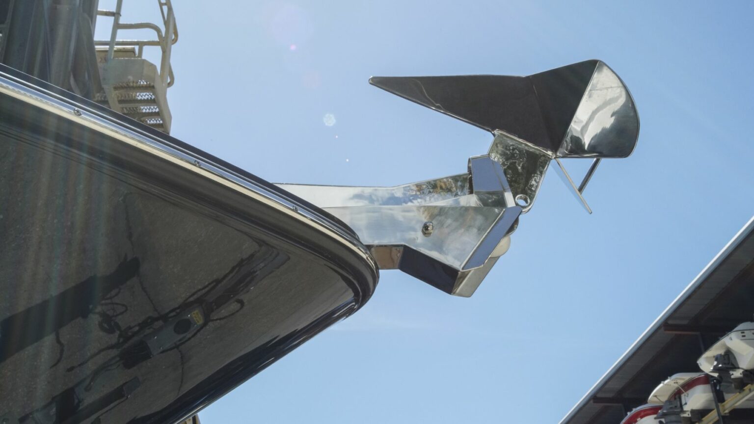 License-free image - A close-up view of a boat&#039;s anchor system mounted at the bow. The polished metallic anchor and mount are contrasted against the clear blue sky, with the sun creating reflections and lens flare. The boat and part of a metal structure are visible in the background. A plow, CQR, wing or delta anchor.