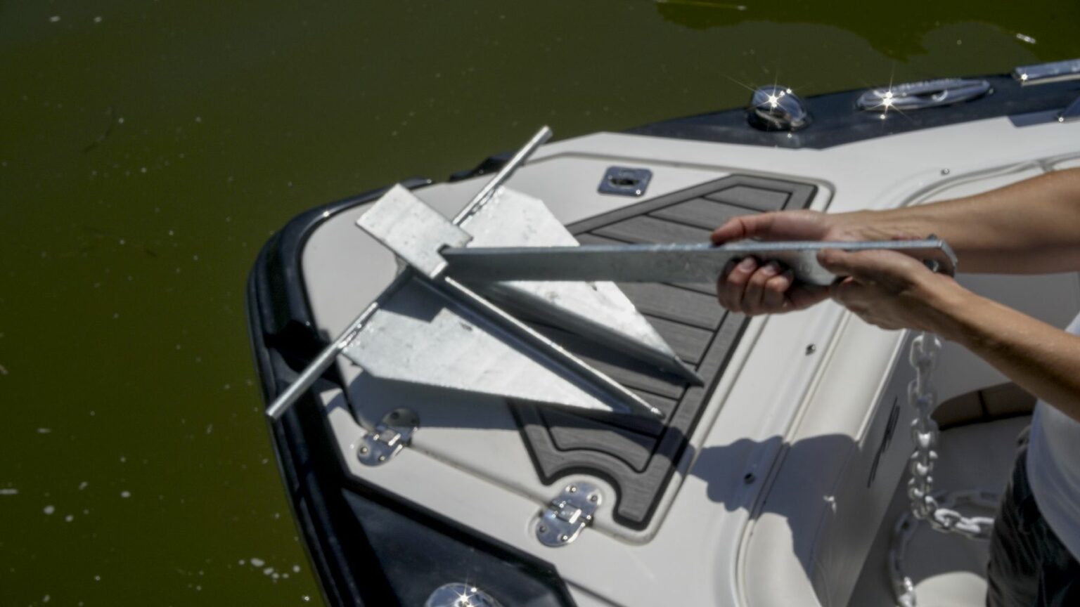 License-free image - A person holding a fluke-style anchor above the deck of a white and black boat. The boat is on a greenish body of water, and metallic boat cleats can be seen on the deck. A fluke, danforth, or lightweight anchor.